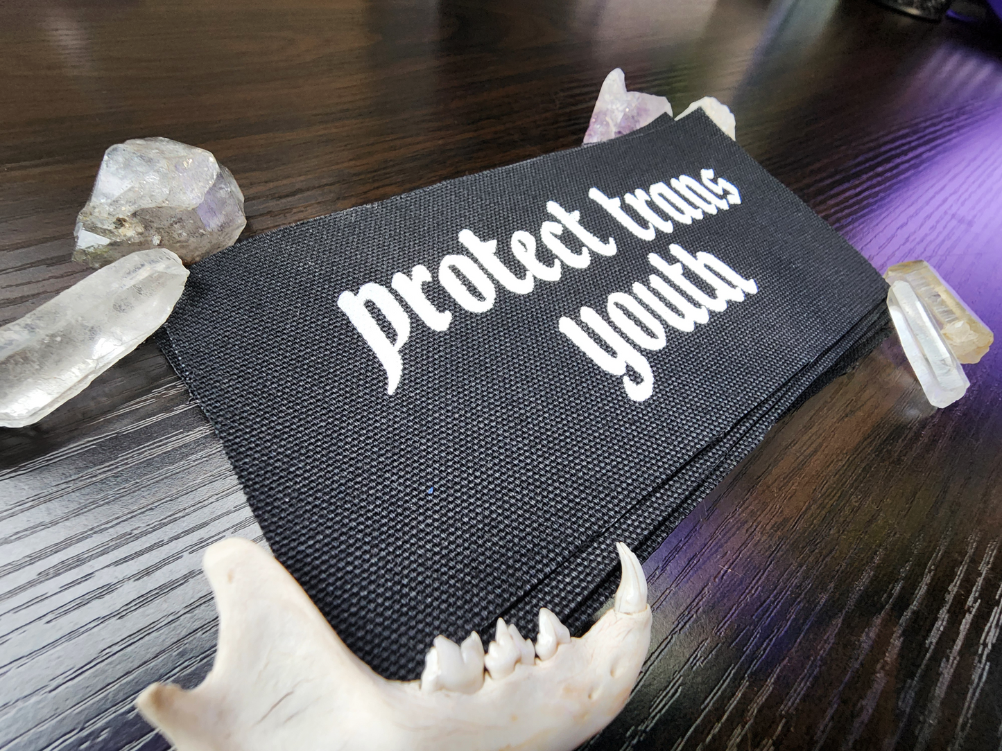 A black fabric patch with the phrase 'Protect Trans Youth' printed in bold, white Gothic-style lettering. The patch is stacked neatly with others on a dark wooden surface. Surrounding the patch are decorative elements, including clear and purple crystals, a small animal jawbone, and rough stones, adding a mystical and earthy aesthetic.