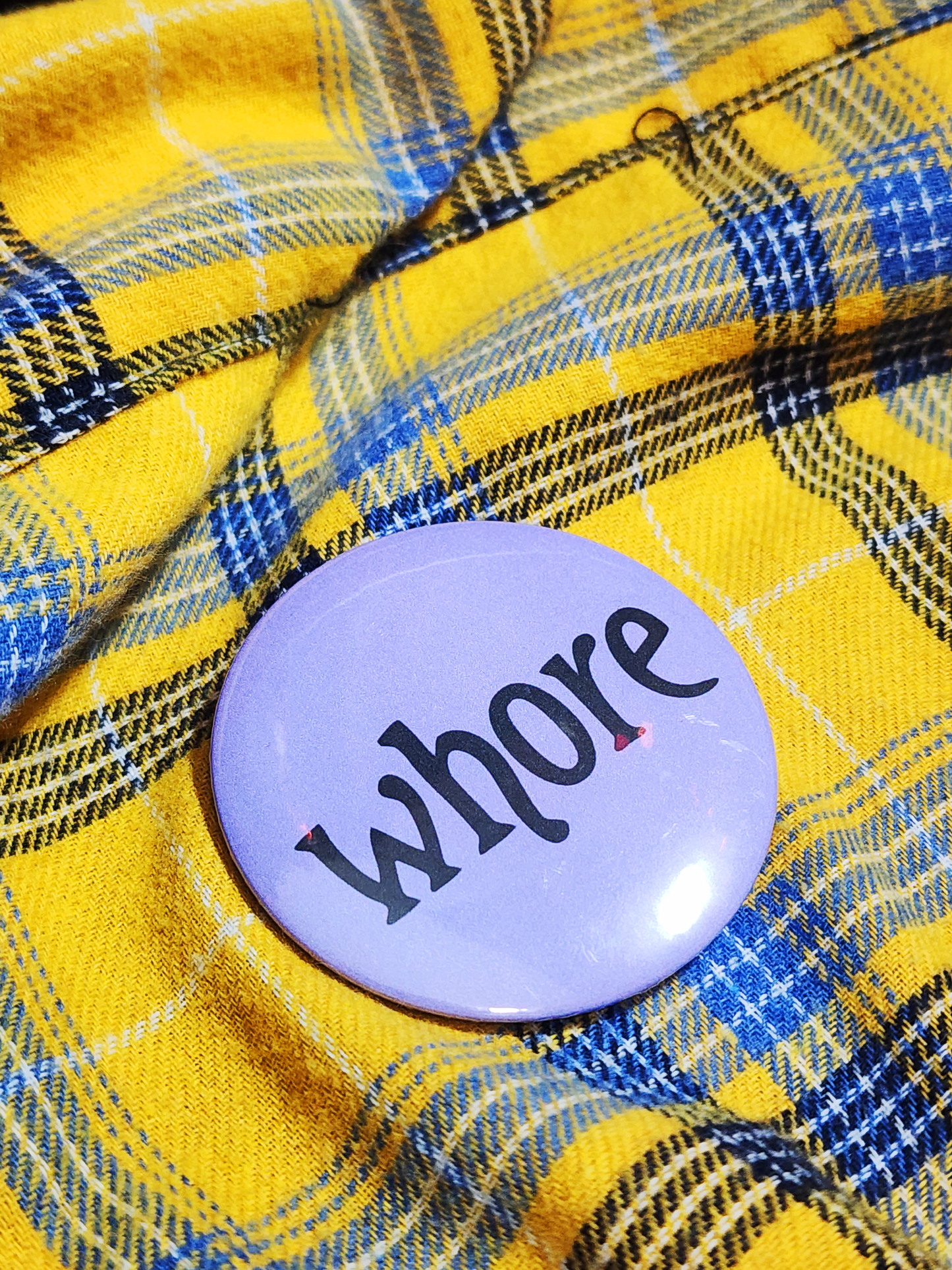 the pin sits on a yellow flannel. the pin is purple with black decorative type. the type reads "whore" in all lowercase letters. the background is pastel purple.