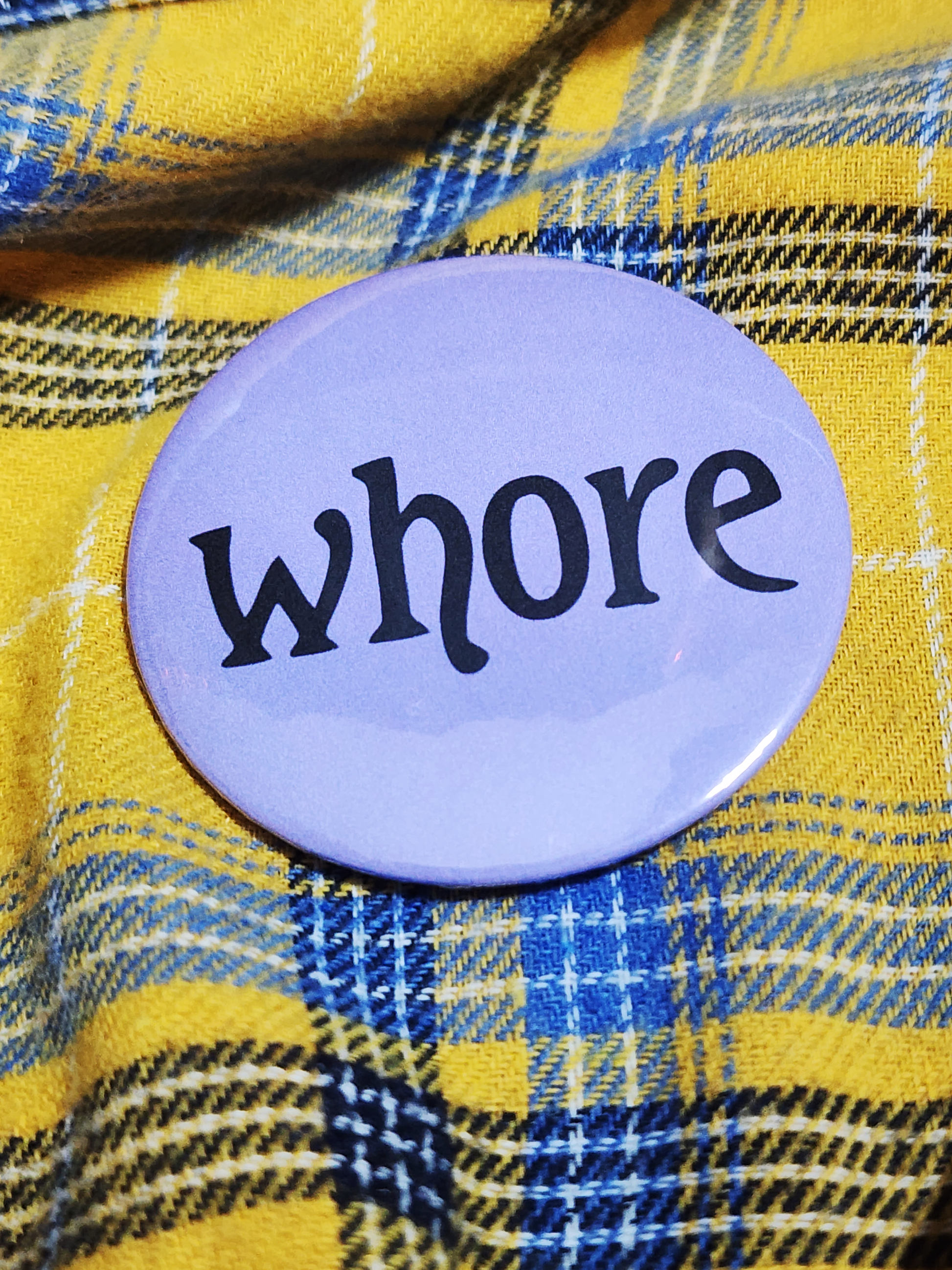 the pin sits on a yellow flannel. the pin is purple with black decorative type. the type reads "whore" in all lowercase letters. the background is pastel purple.
