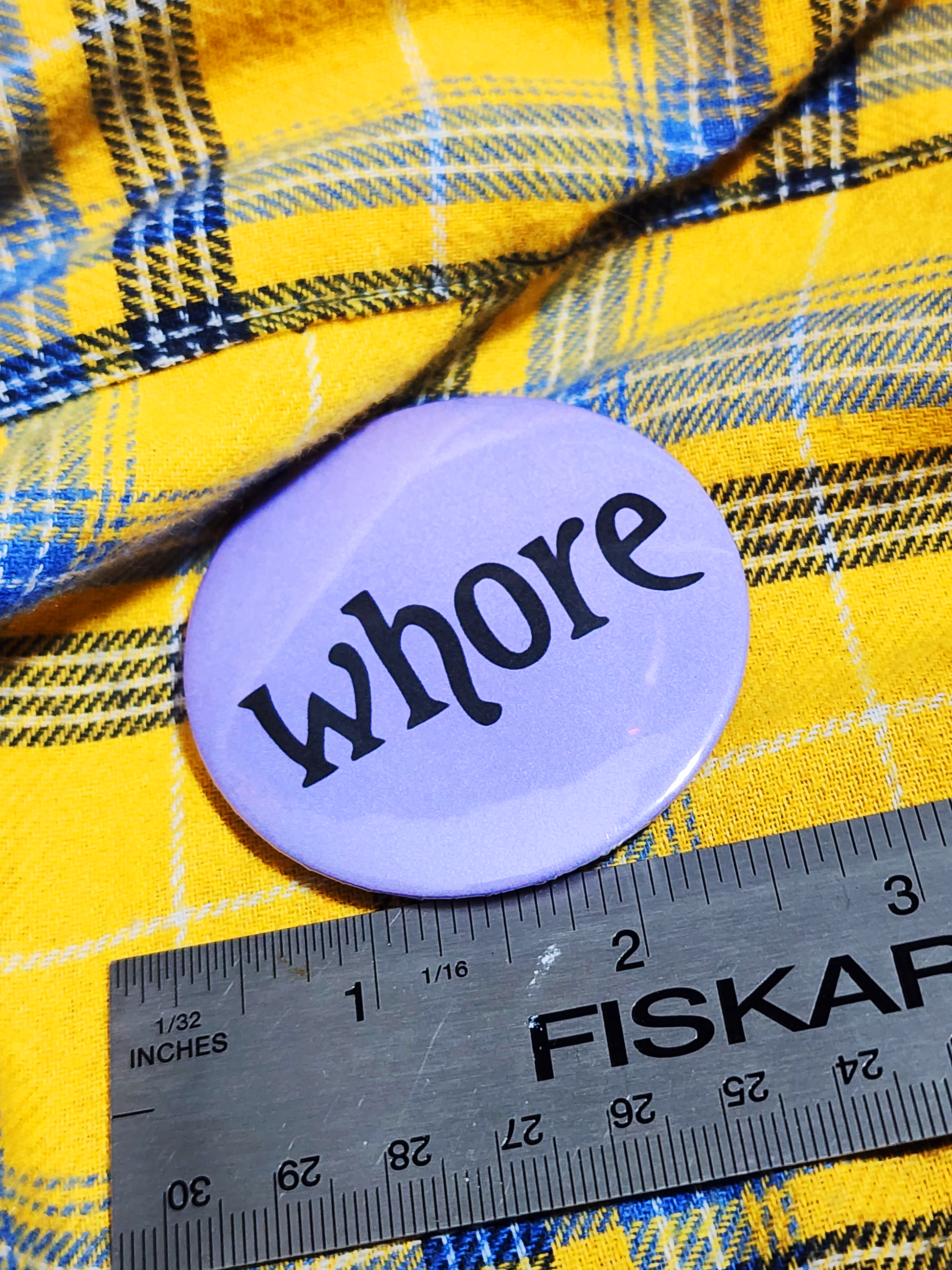 the pin sits on a yellow flannel. the pin is purple with black decorative type. the type reads "whore" in all lowercase letters. the background is pastel purple.