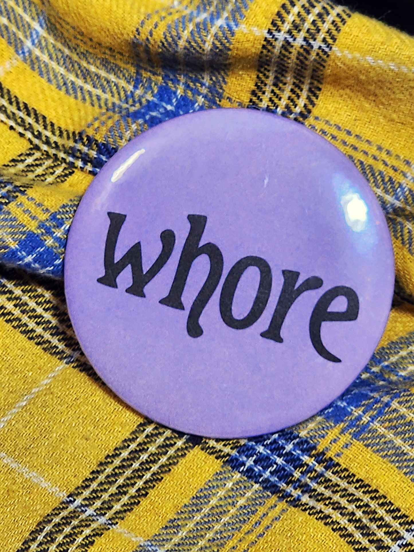 the pin sits on a yellow flannel. the pin is purple with black decorative type. the type reads "whore" in all lowercase letters. the background is pastel purple.