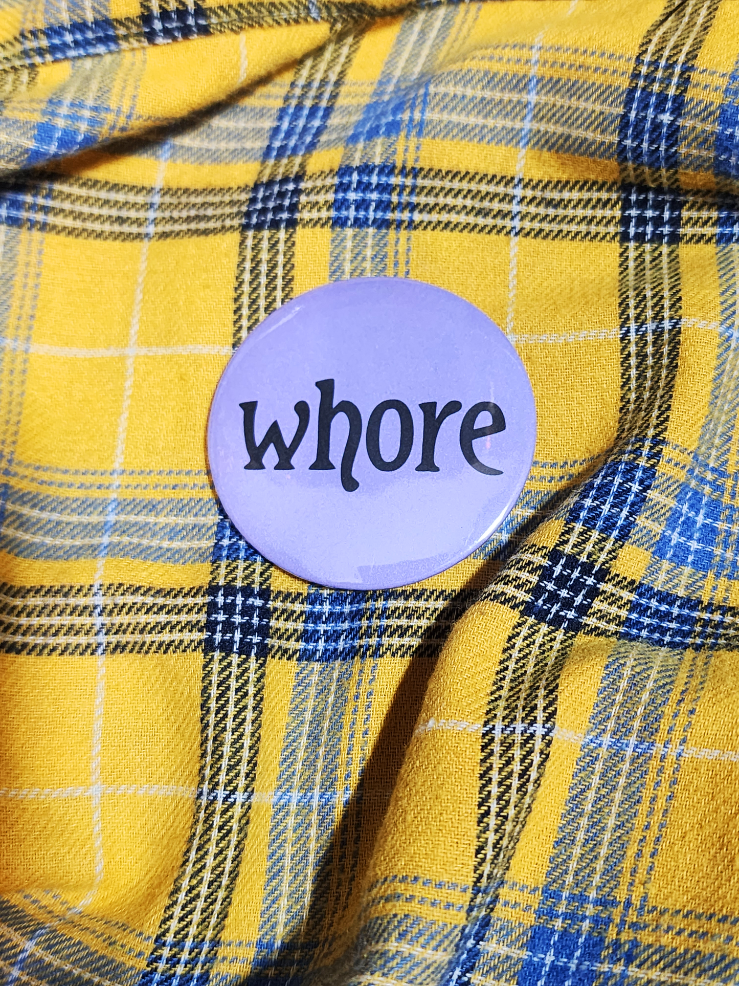 a hand with visible tattoos and painted nails holds the pin against the yellow flannel surface. the pin is purple with black decorative type. the type reads "whore" in all lowercase letters. the background of the pin is pastel purple.