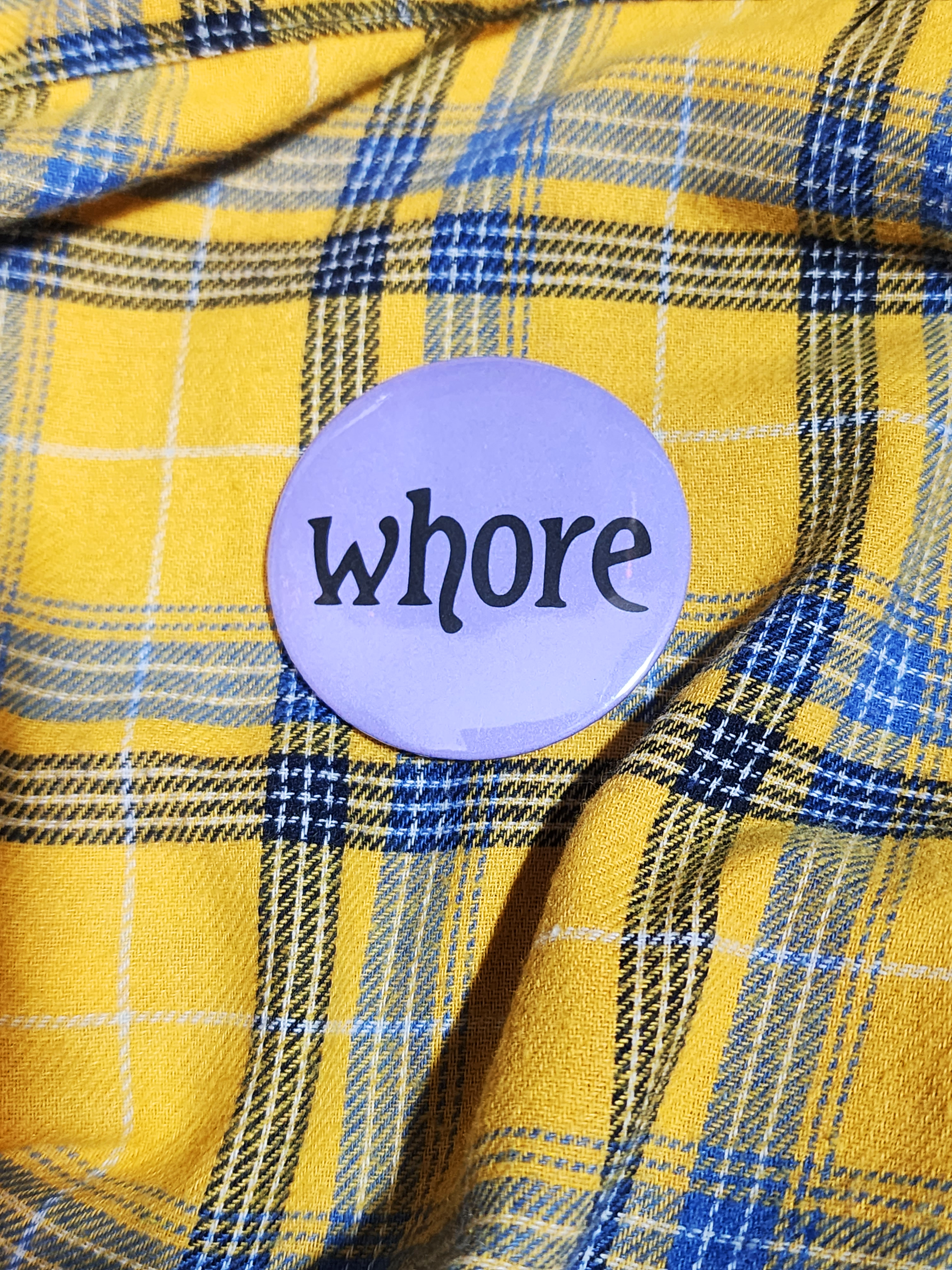 a hand with visible tattoos and painted nails holds the pin against the yellow flannel surface. the pin is purple with black decorative type. the type reads "whore" in all lowercase letters. the background of the pin is pastel purple.