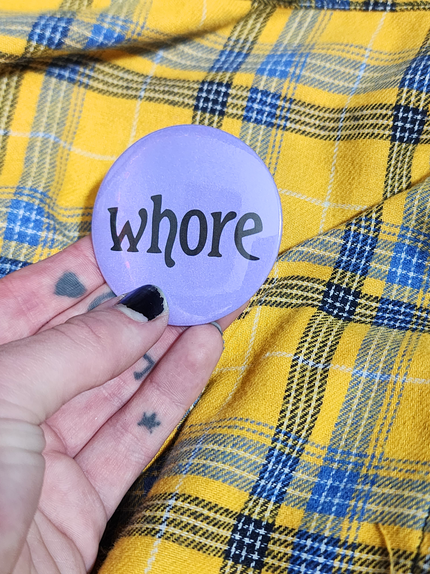 a hand with visible tattoos and painted nails holds the pin against the yellow flannel surface. the pin is purple with black decorative type. the type reads "whore" in all lowercase letters. the background of the pin is pastel purple.