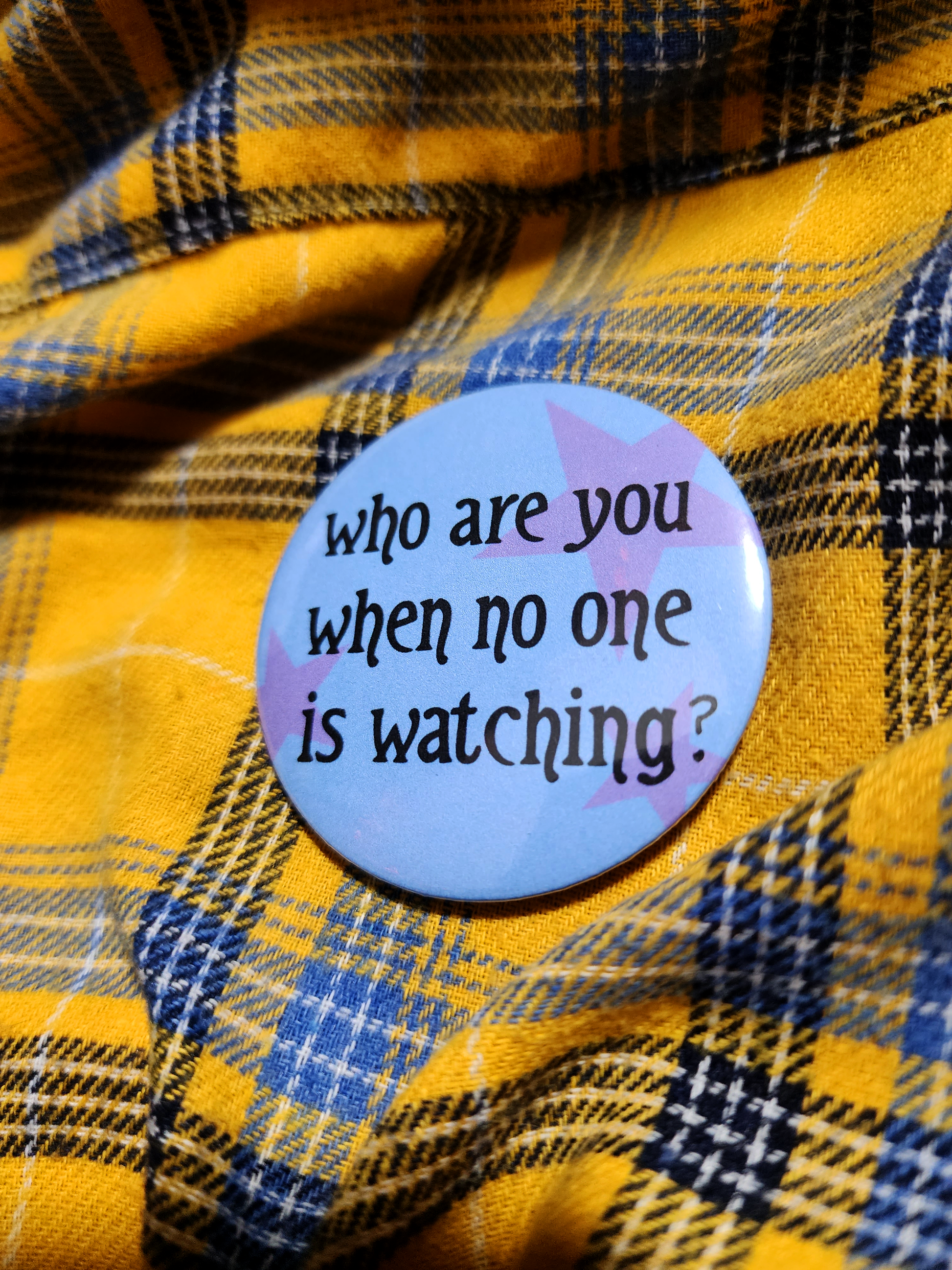 the pin sits on a yellow flannel. the pin is blue with purple decorative stars in the background. the black type reads "who are you when no one is watching?" in all lowercase letters.