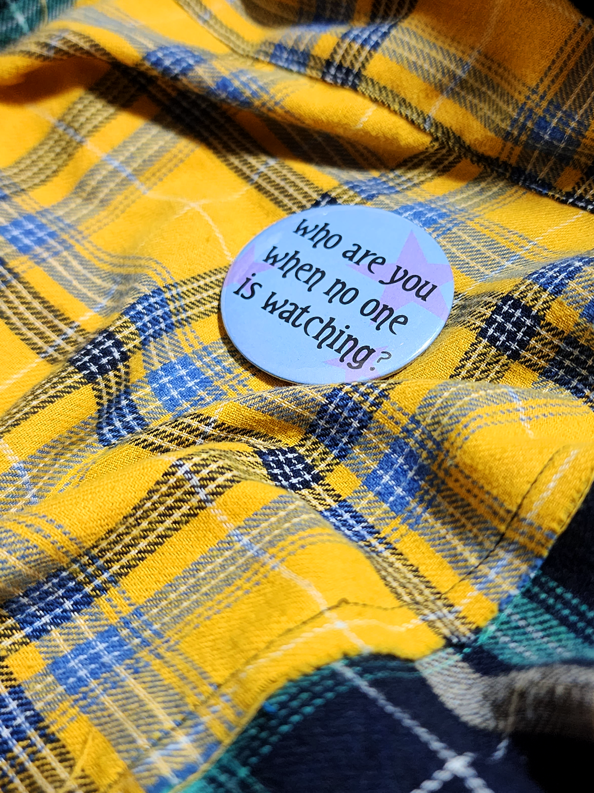 the pin sits on a yellow flannel. the pin is blue with purple decorative stars in the background. the black type reads "who are you when no one is watching?" in all lowercase letters.
