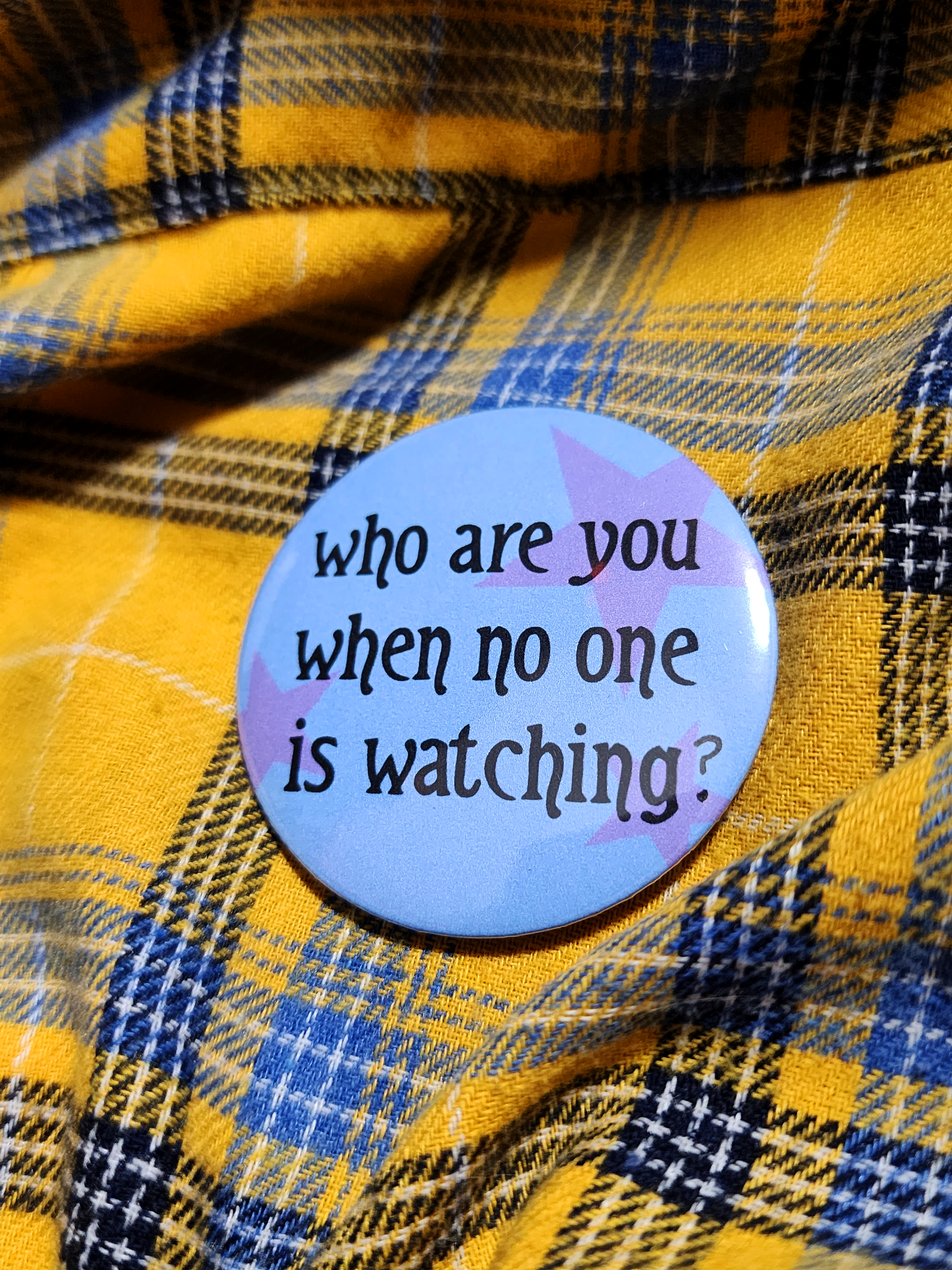 the pin sits on a yellow flannel. the pin is blue with purple decorative stars in the background. the black type reads "who are you when no one is watching?" in all lowercase letters.