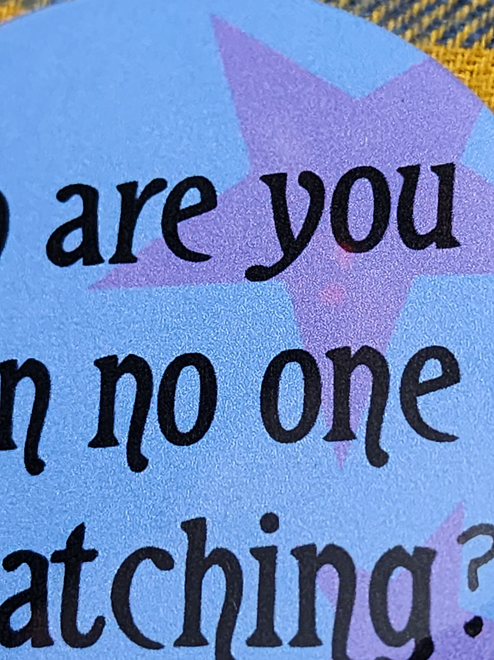 the pin sits on a yellow flannel. the pin is blue with purple decorative stars in the background. the black type reads "who are you when no one is watching?" in all lowercase letters.