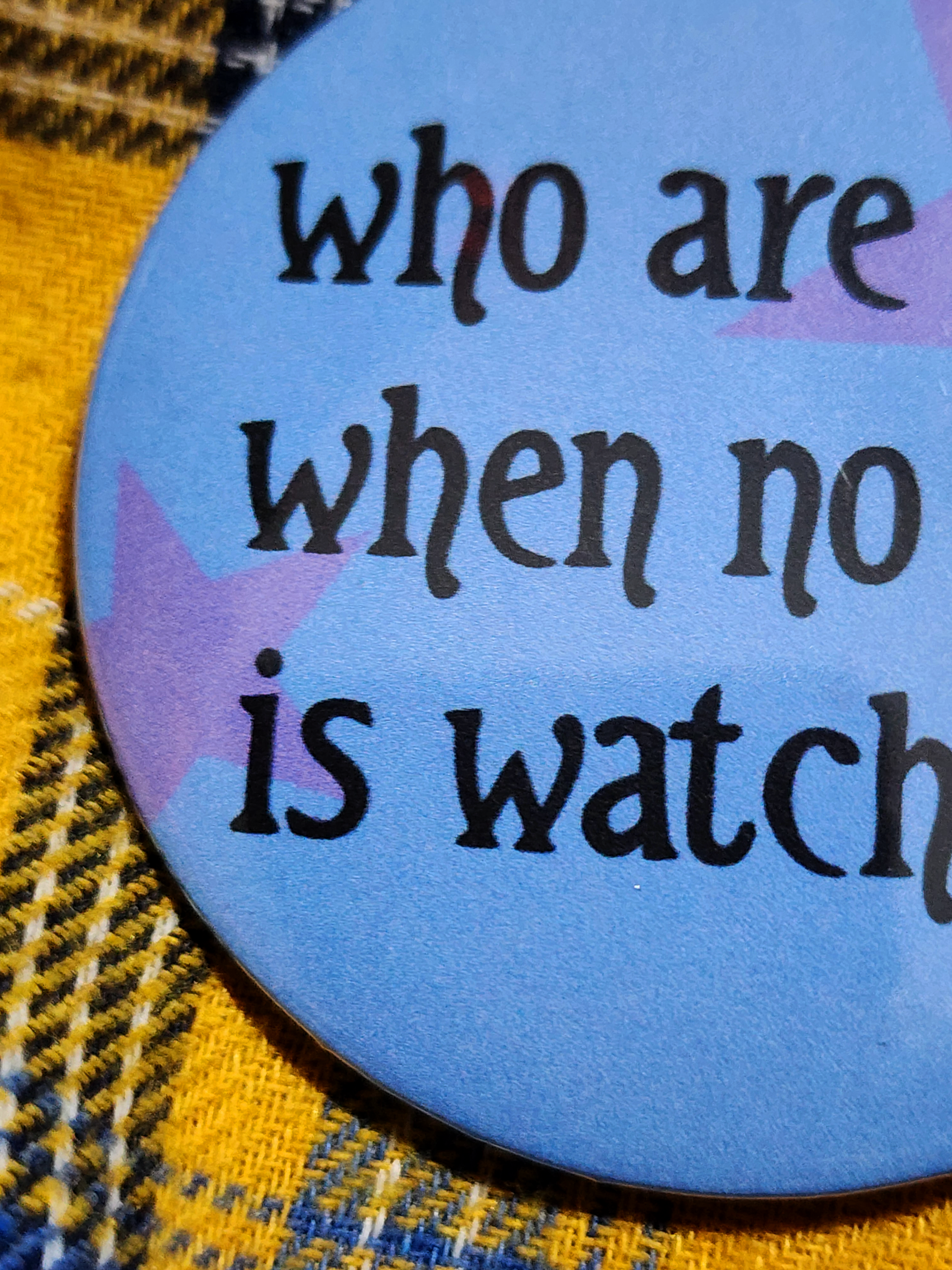 the pin sits on a yellow flannel. the pin is blue with purple decorative stars in the background. the black type reads "who are you when no one is watching?" in all lowercase letters.