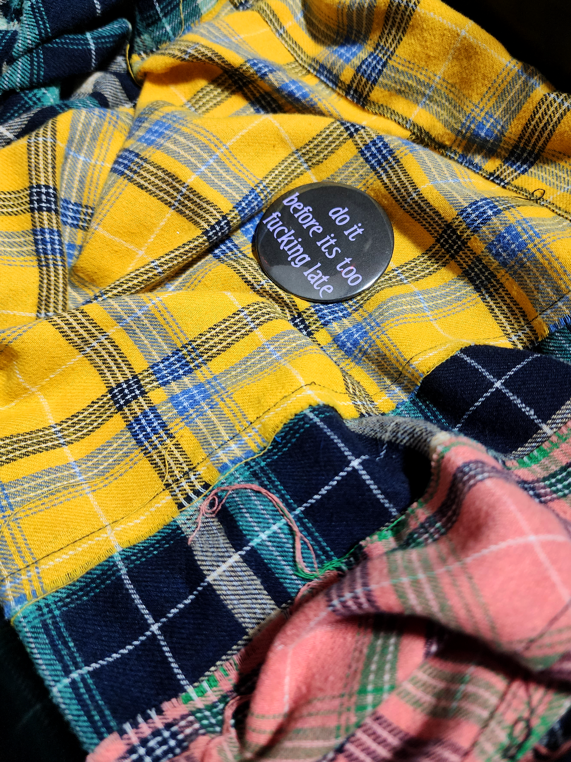 the pin sits on a yellow flannel. the pin is black with purple decorative type. the type reads "do it before it's too fucking late" in all lowercase letters.