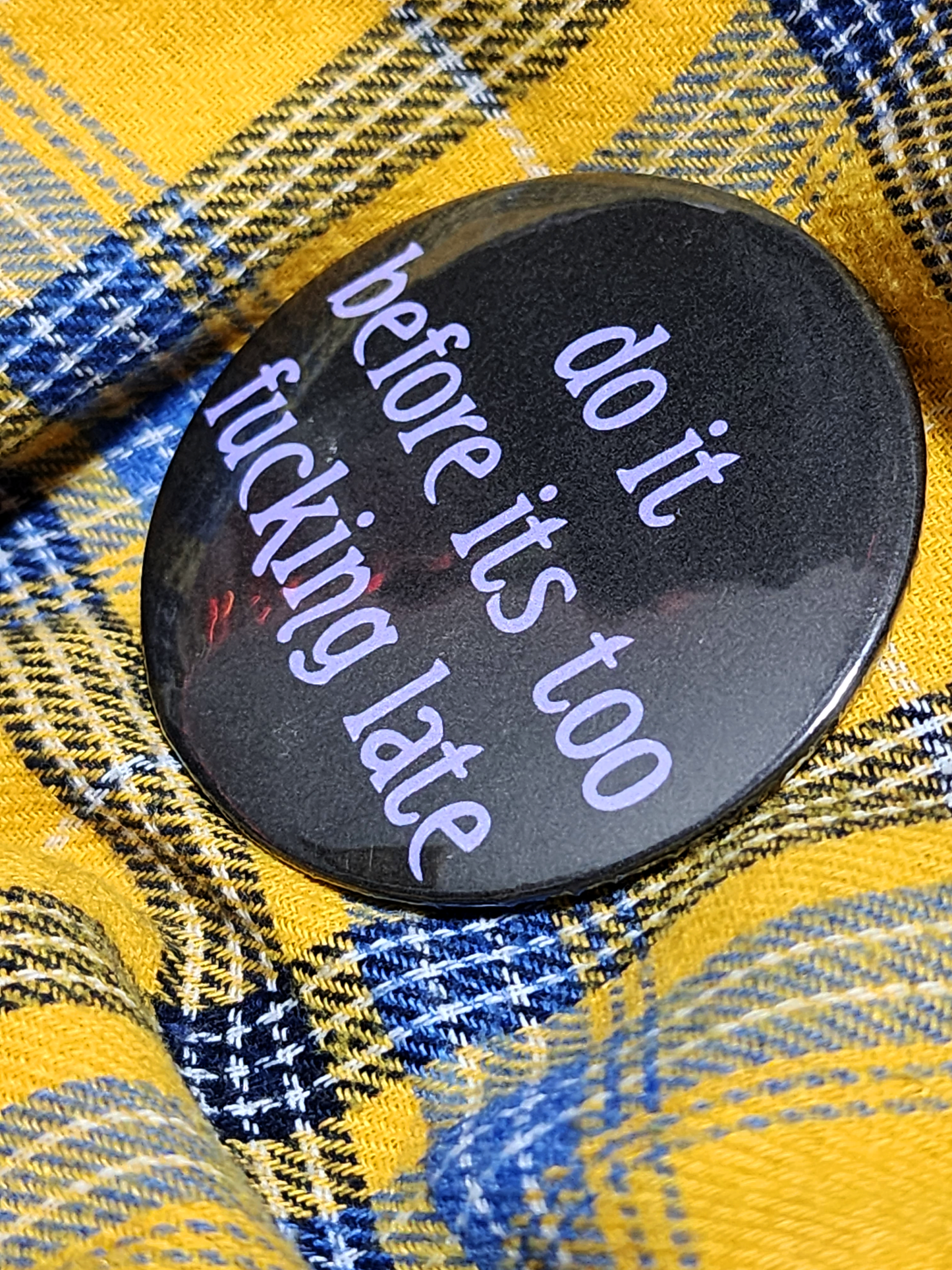 the pin sits on a yellow flannel. the pin is black with purple decorative type. the type reads "do it before it's too fucking late" in all lowercase letters.