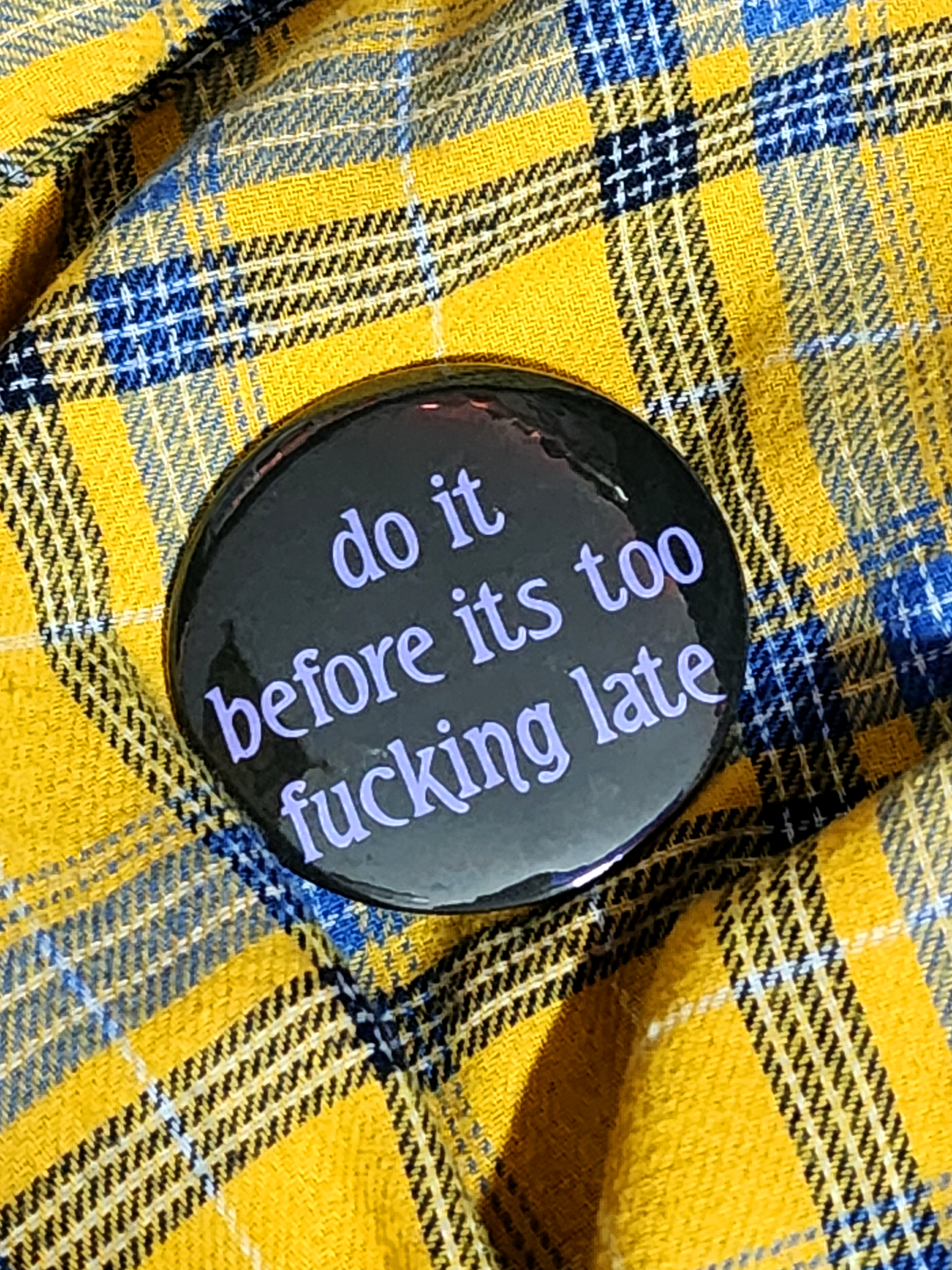 the pin sits on a yellow flannel. the pin is black with purple decorative type. the type reads "do it before it's too fucking late" in all lowercase letters.