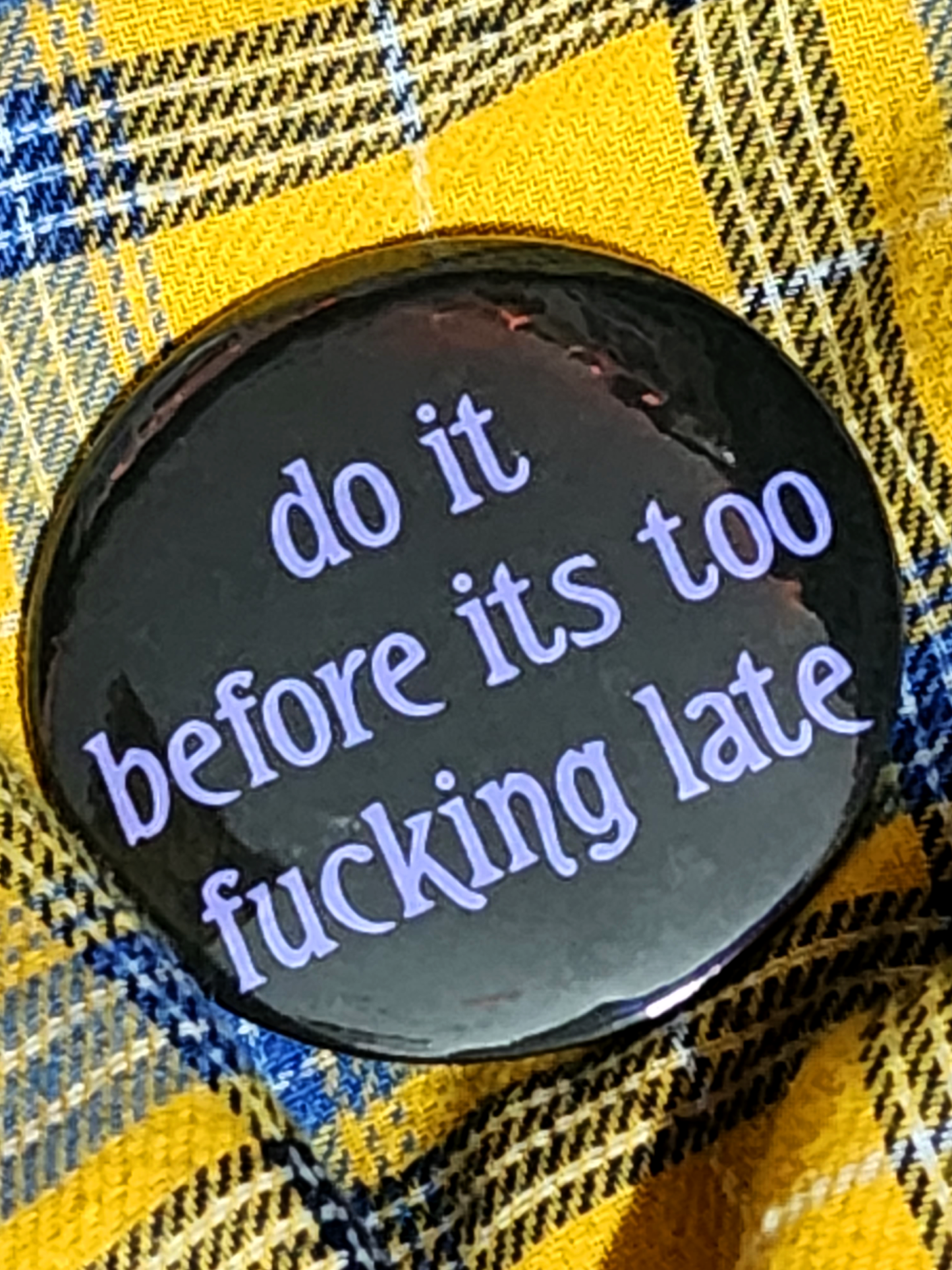 the pin sits on a yellow flannel. the pin is black with purple decorative type. the type reads "do it before it's too fucking late" in all lowercase letters.