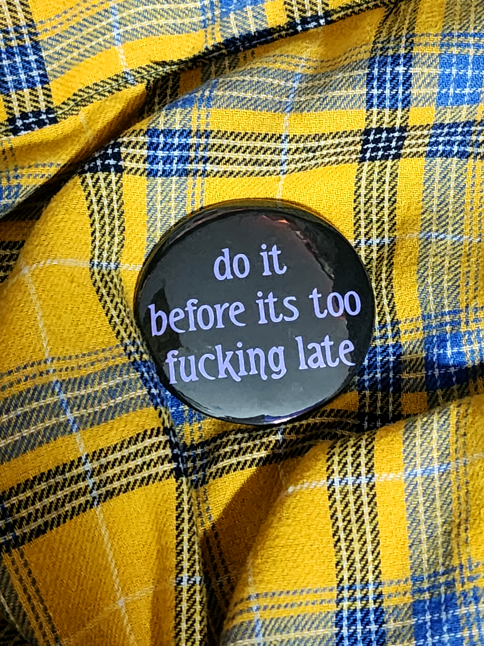the pin sits on a yellow flannel. the pin is black with purple decorative type. the type reads "do it before it's too fucking late" in all lowercase letters.