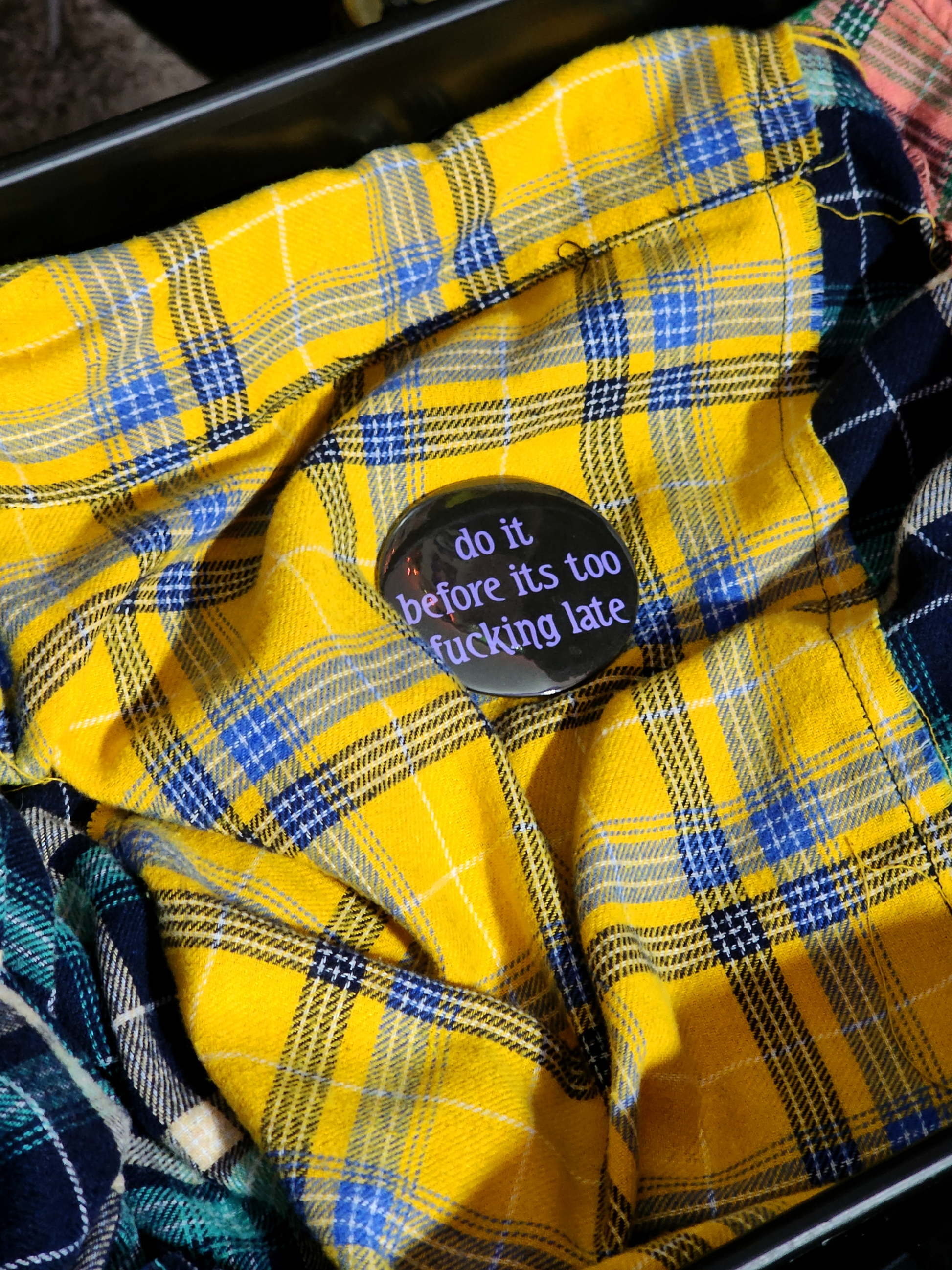 the pin sits on a yellow flannel. the pin is black with purple decorative type. the type reads "do it before it's too fucking late" in all lowercase letters.