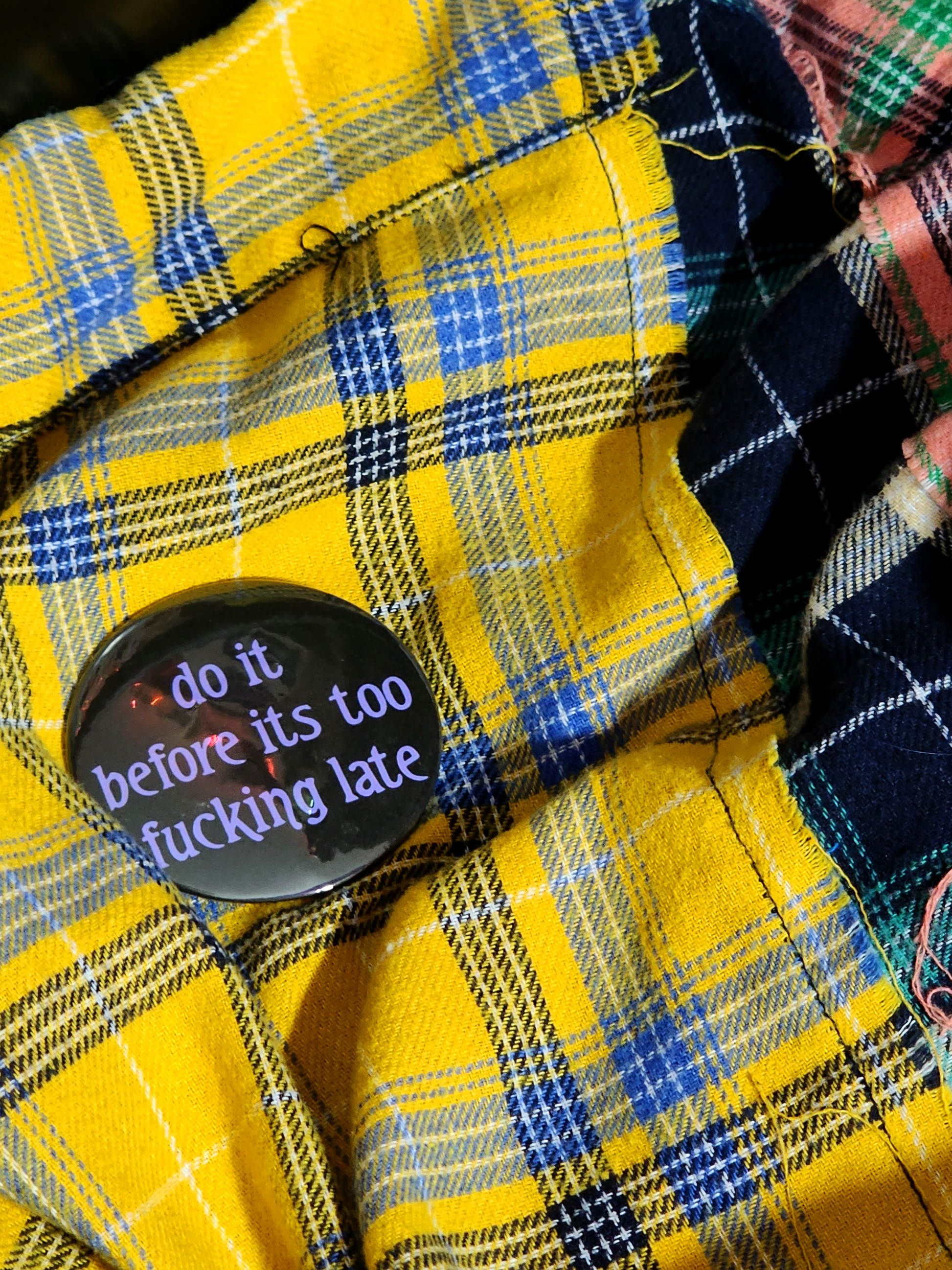 the pin sits on a yellow flannel. the pin is black with purple decorative type. the type reads "do it before it's too fucking late" in all lowercase letters.