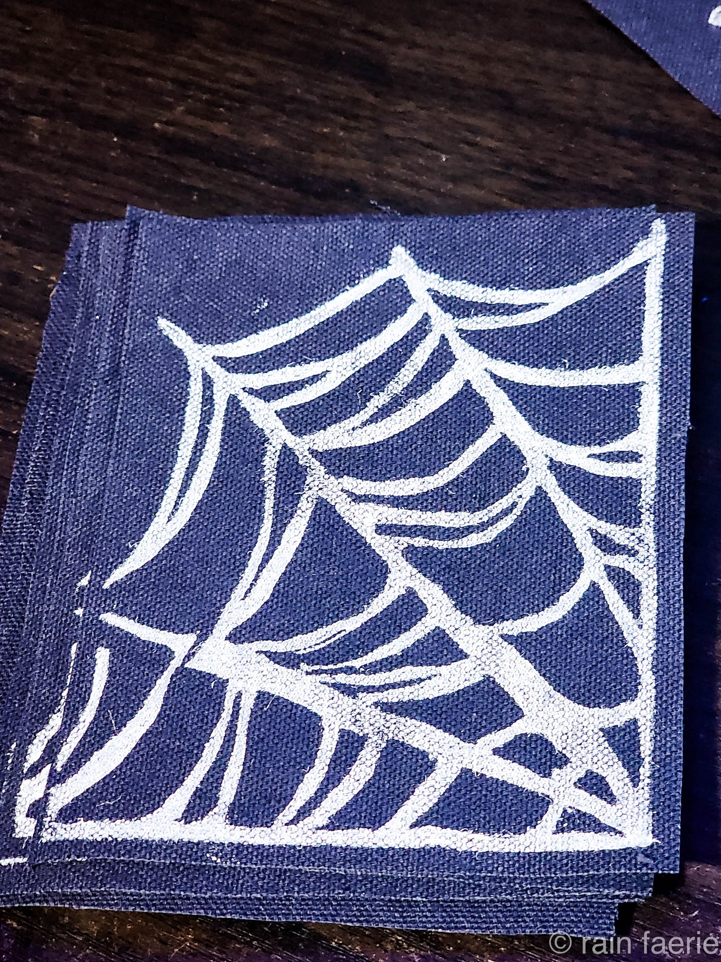 Stack of black fabric patches with a bold white linocut design of a spiderweb, printed by hand. The patches are placed on a dark wooden surface. Perfect for sewing onto crust pants, battle vests, battle jackets, or any fabric surface!!
