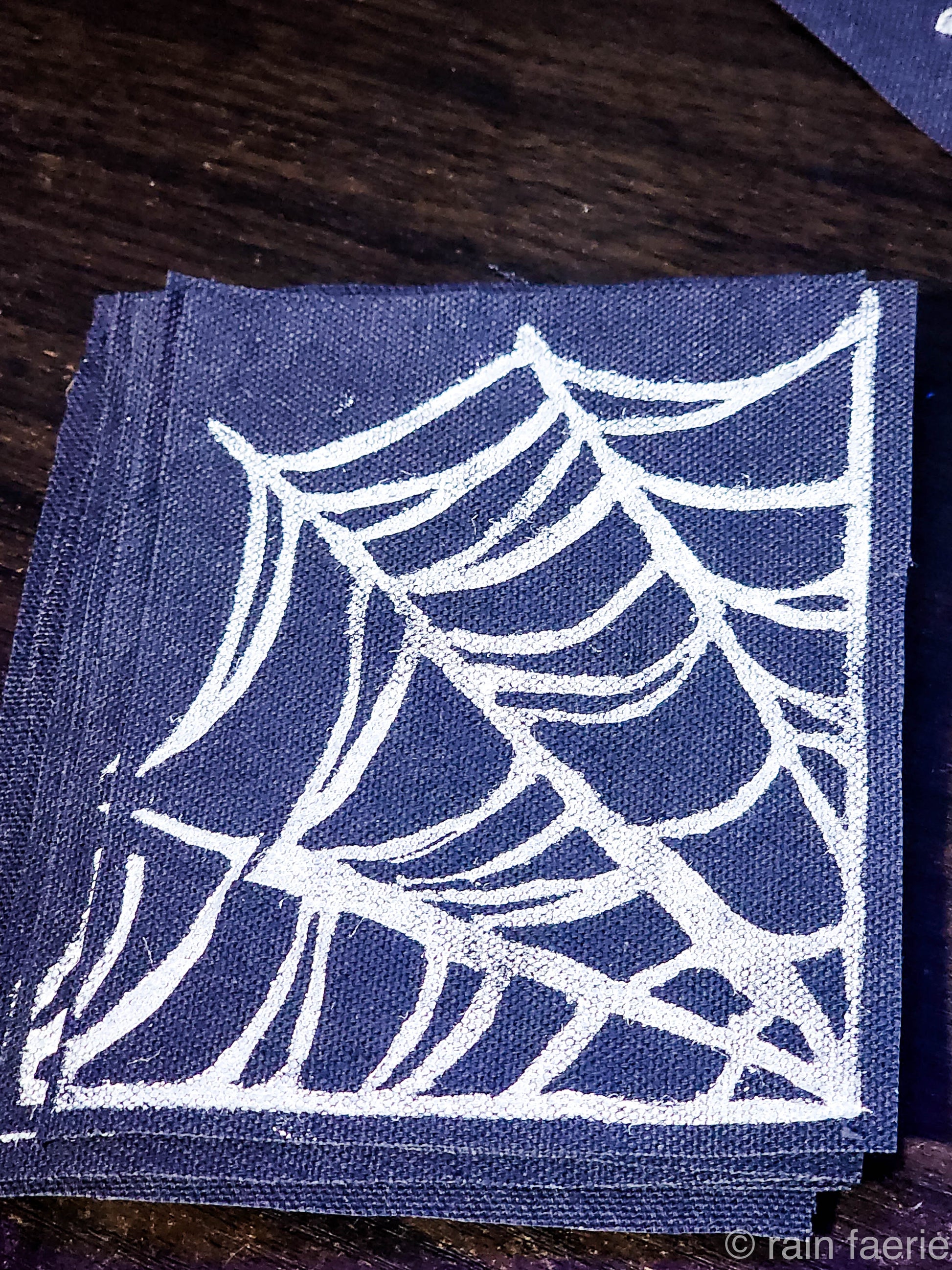 Stack of black fabric patches with a bold white linocut design of a spiderweb, printed by hand. The patches are placed on a dark wooden surface. Perfect for sewing onto crust pants, battle vests, battle jackets, or any fabric surface!!