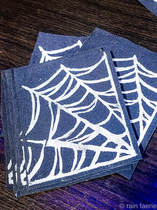 Stack of black fabric patches with a bold white linocut design of a spiderweb, printed by hand. The patches are placed on a dark wooden surface. Perfect for sewing onto crust pants, battle vests, battle jackets, or any fabric surface!!