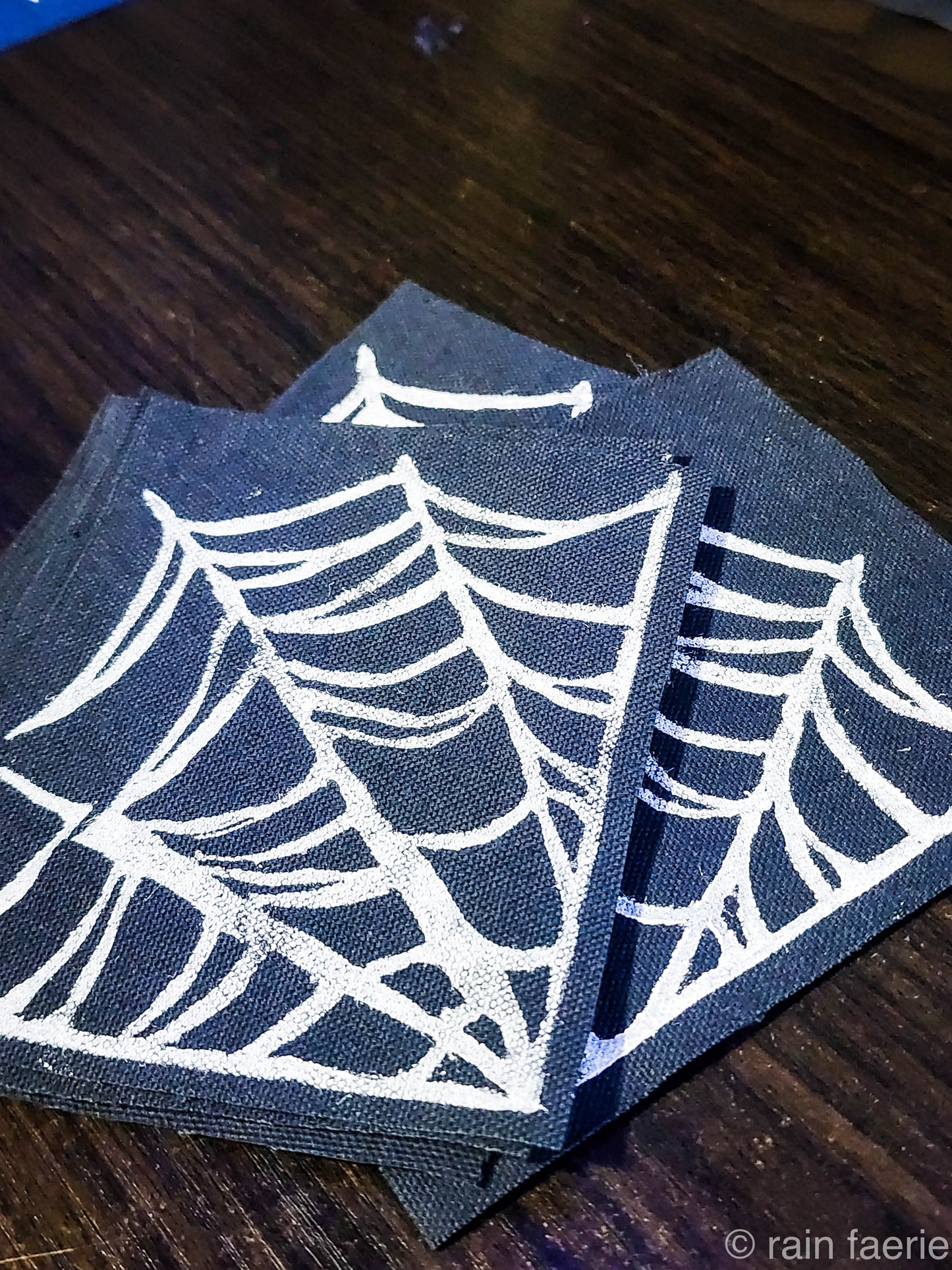 Stack of black fabric patches with a bold white linocut design of a spiderweb, printed by hand. The patches are placed on a dark wooden surface. Perfect for sewing onto crust pants, battle vests, battle jackets, or any fabric surface!!