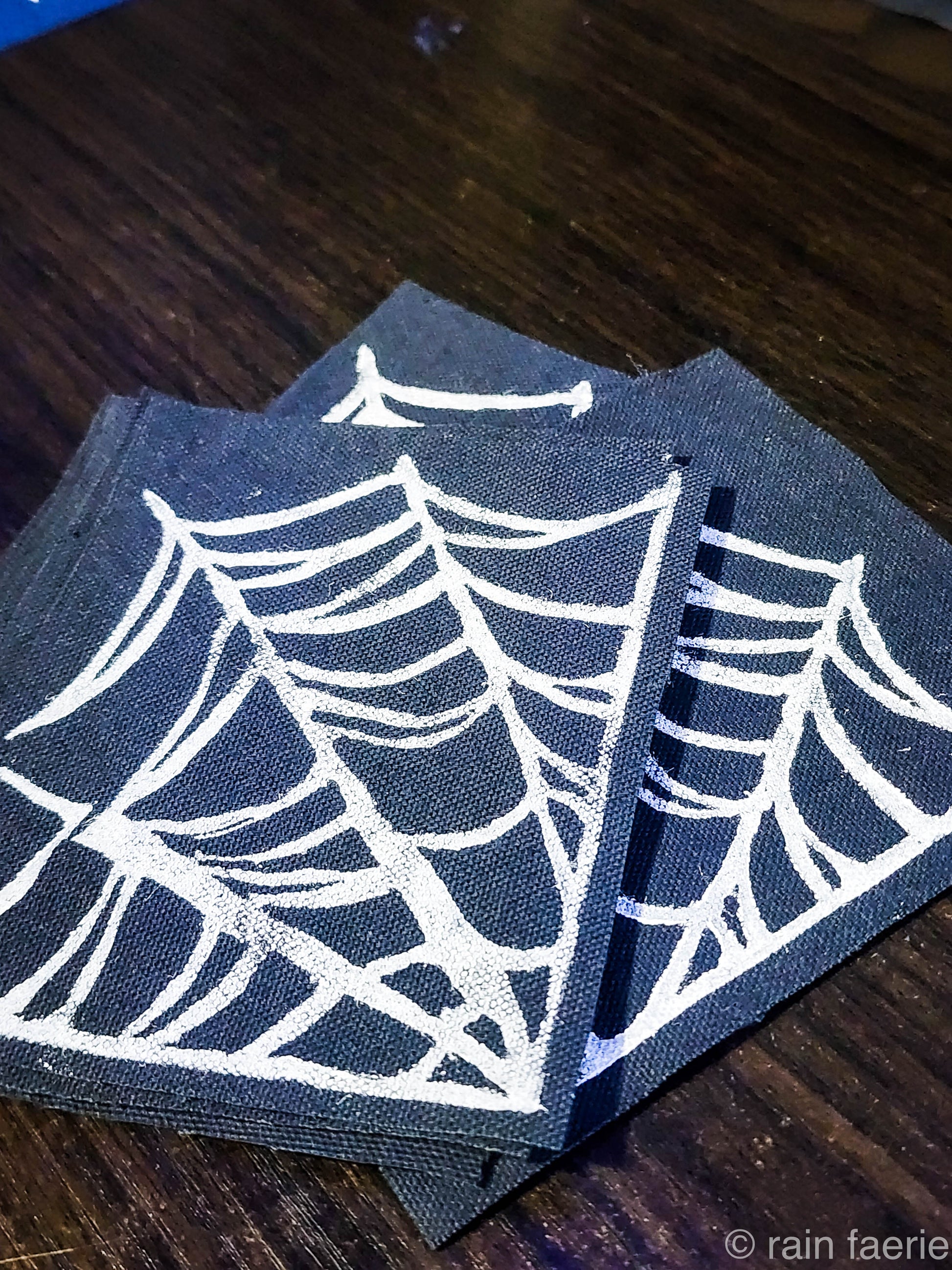 Stack of black fabric patches with a bold white linocut design of a spiderweb, printed by hand. The patches are placed on a dark wooden surface. Perfect for sewing onto crust pants, battle vests, battle jackets, or any fabric surface!!