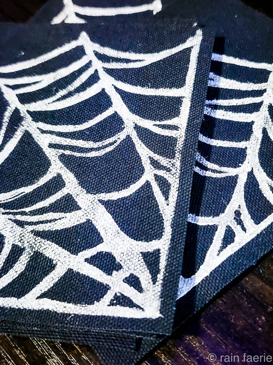 Stack of black fabric patches with a bold white linocut design of a spiderweb, printed by hand. The patches are placed on a dark wooden surface. Perfect for sewing onto crust pants, battle vests, battle jackets, or any fabric surface!!
