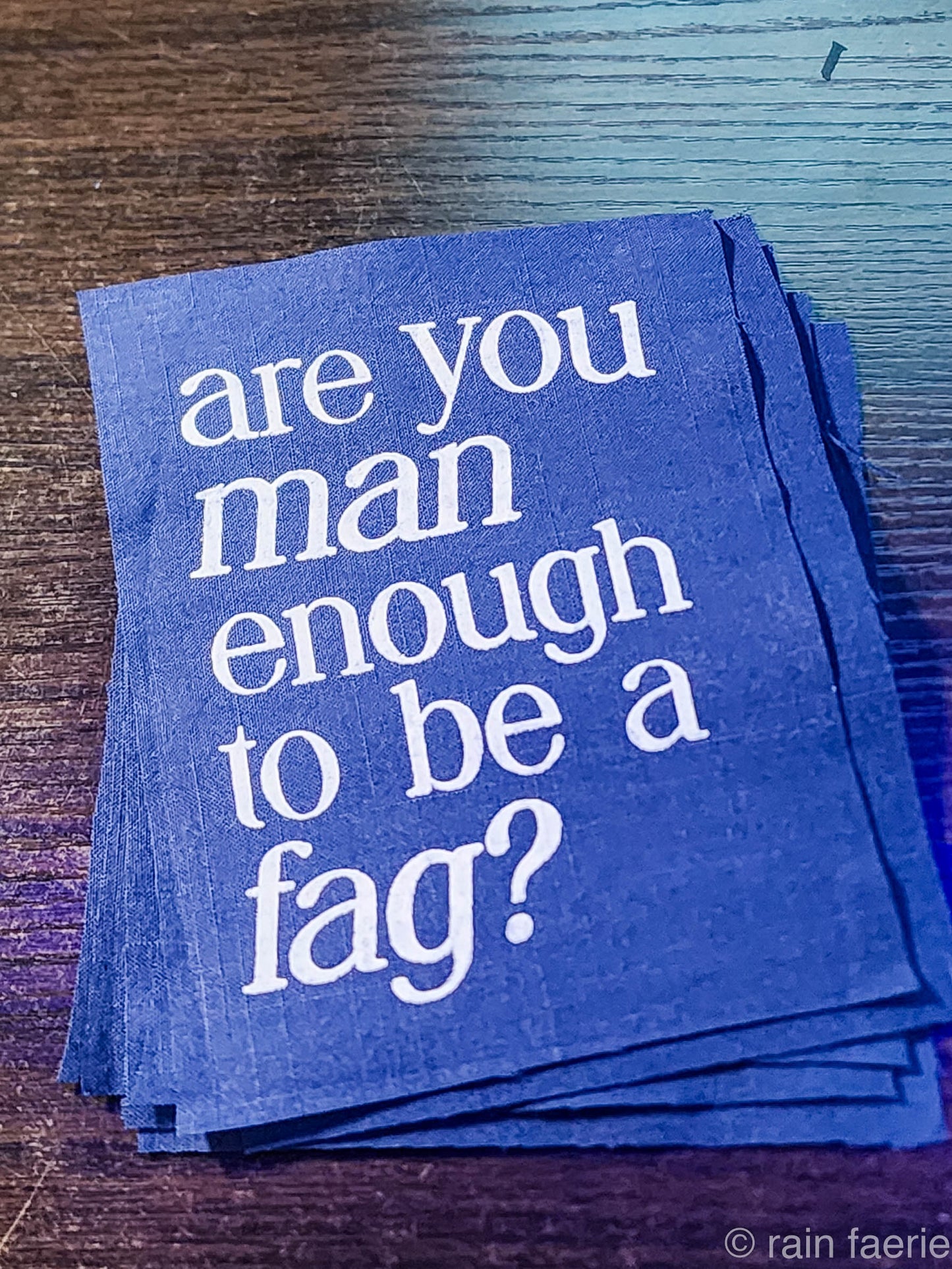 a stack of dark blue patches with bold white paint on a dark wooden surface. lettering is very clean, and a serif font, all lowercase. the patch reads "are you man enough to be a fag?"