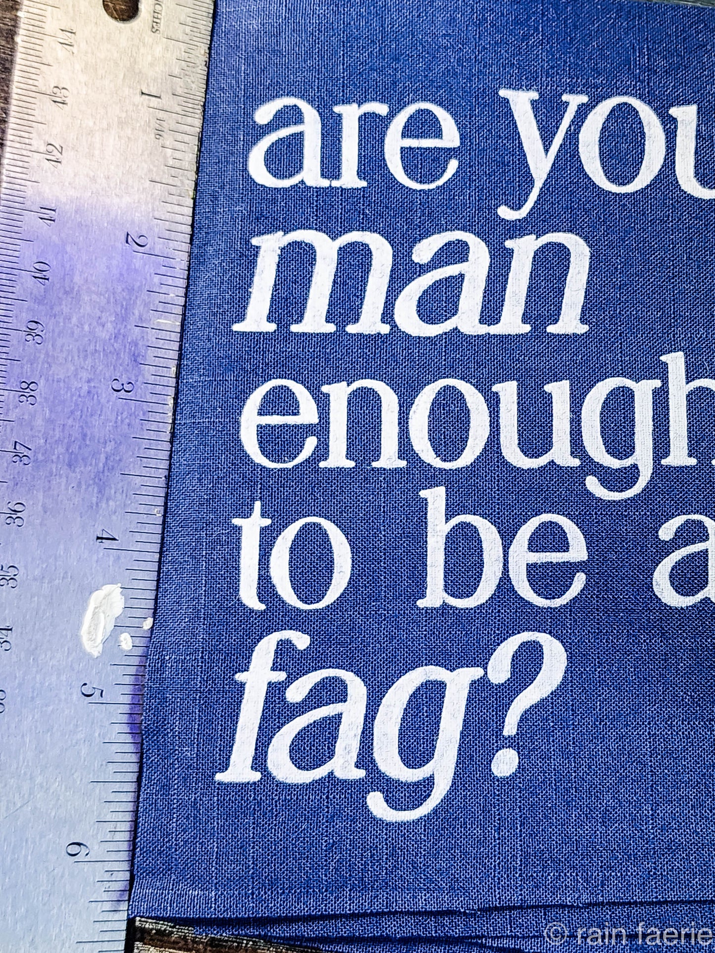 a stack of dark blue patches with bold white paint on a dark wooden surface. lettering is very clean, and a serif font, all lowercase. the patch reads "are you man enough to be a fag?"