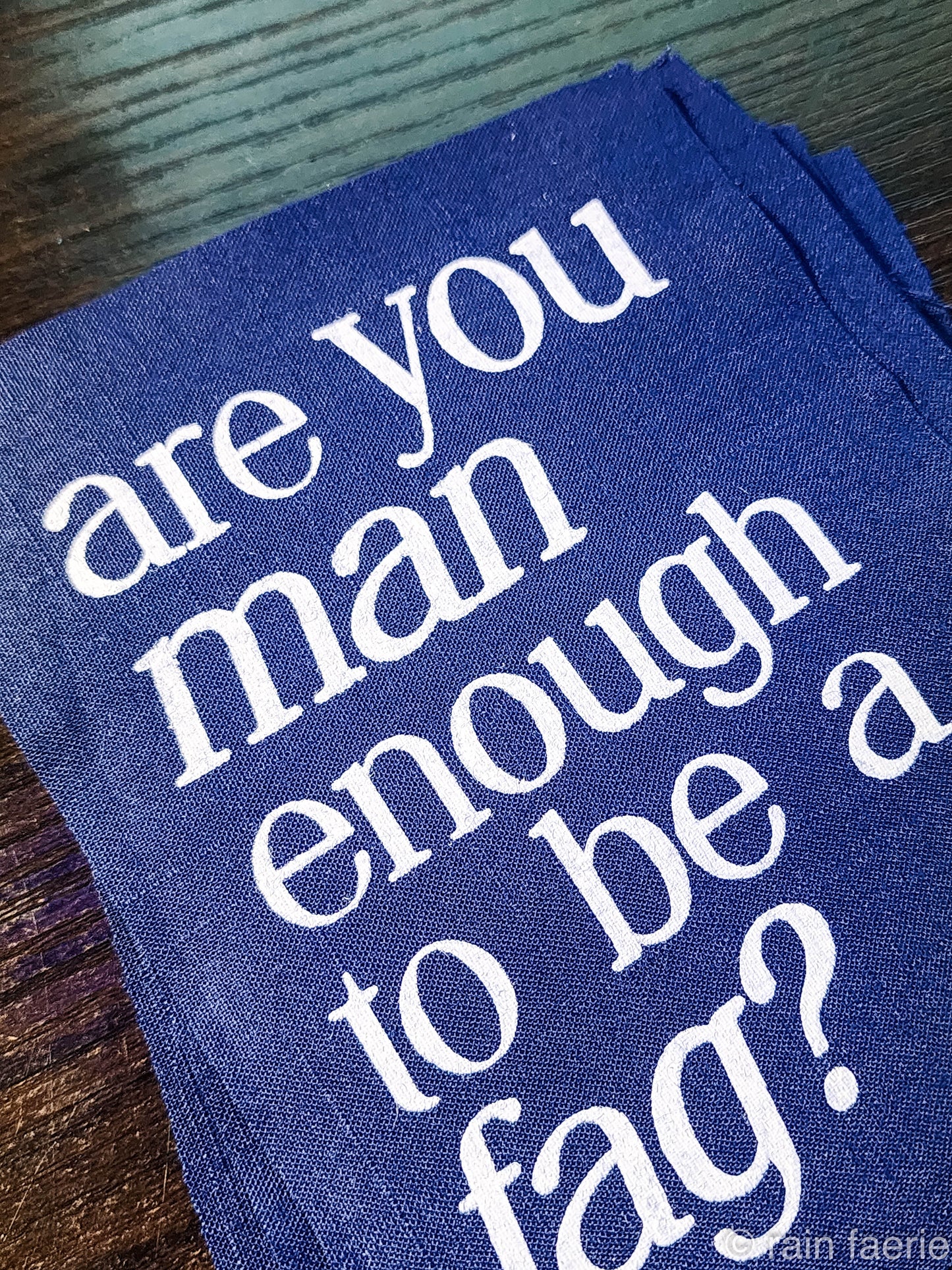 a stack of dark blue patches with bold white paint on a dark wooden surface. lettering is very clean, and a serif font, all lowercase. the patch reads "are you man enough to be a fag?"