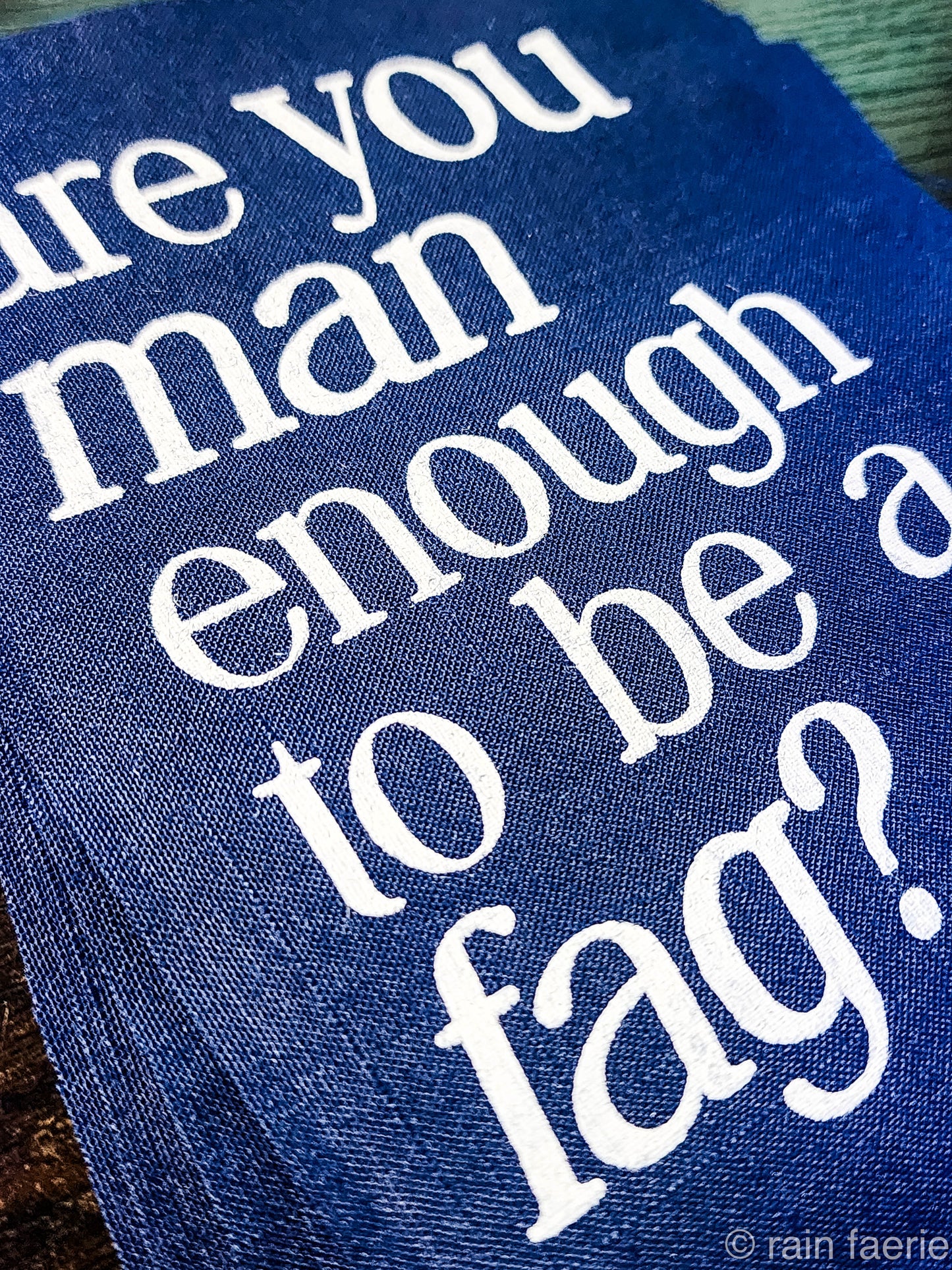 a stack of dark blue patches with bold white paint on a dark wooden surface. lettering is very clean, and a serif font, all lowercase. the patch reads "are you man enough to be a fag?"