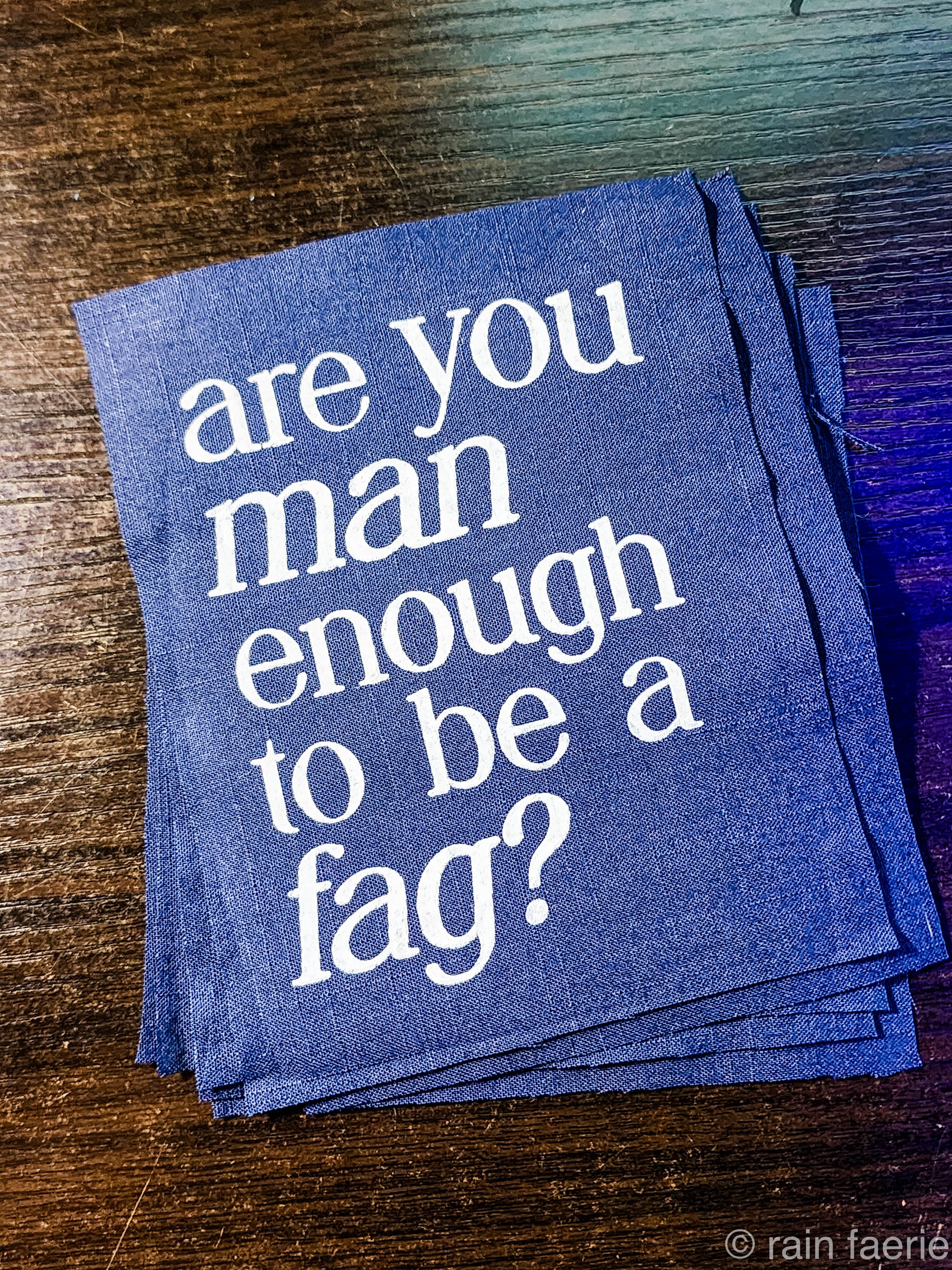 a stack of dark blue patches with bold white paint on a dark wooden surface. lettering is very clean, and a serif font, all lowercase. the patch reads "are you man enough to be a fag?"