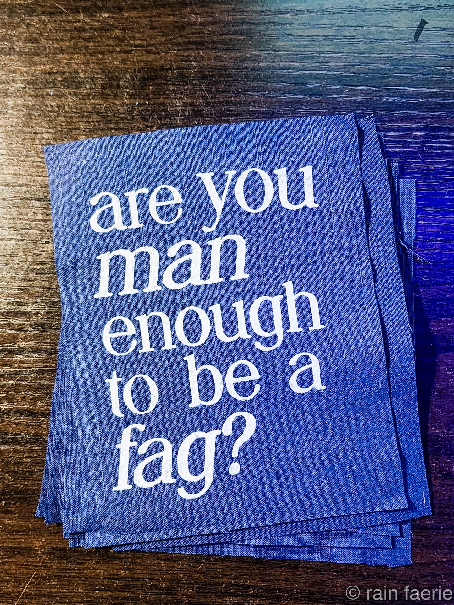 a stack of dark blue patches with bold white paint on a dark wooden surface. lettering is very clean, and a serif font, all lowercase. the patch reads "are you man enough to be a fag?"
