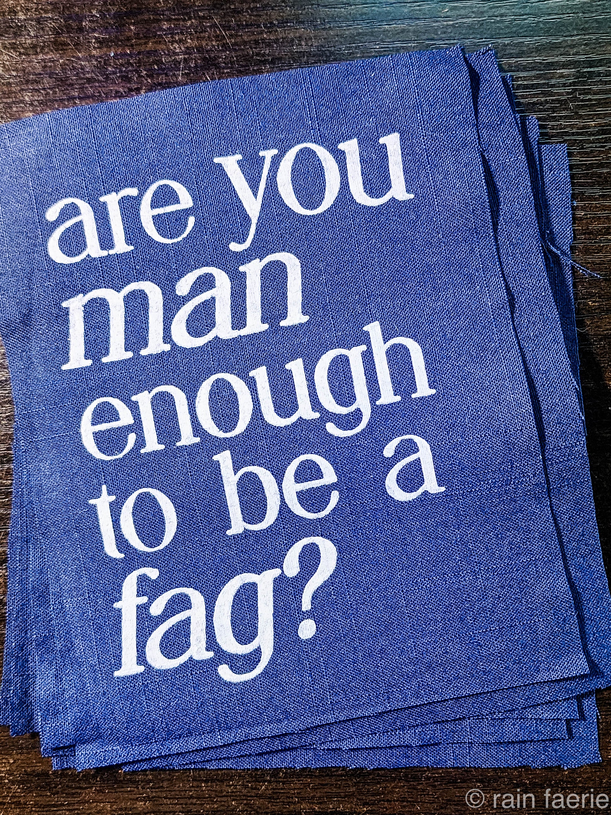 a stack of dark blue patches with bold white paint on a dark wooden surface. lettering is very clean, and a serif font, all lowercase. the patch reads "are you man enough to be a fag?"