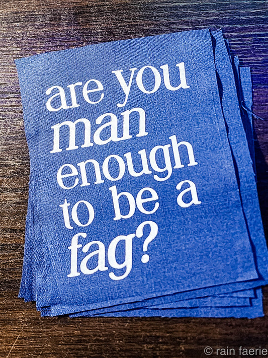 a stack of dark blue patches with bold white paint on a dark wooden surface. lettering is very clean, and a serif font, all lowercase. the patch reads "are you man enough to be a fag?"
