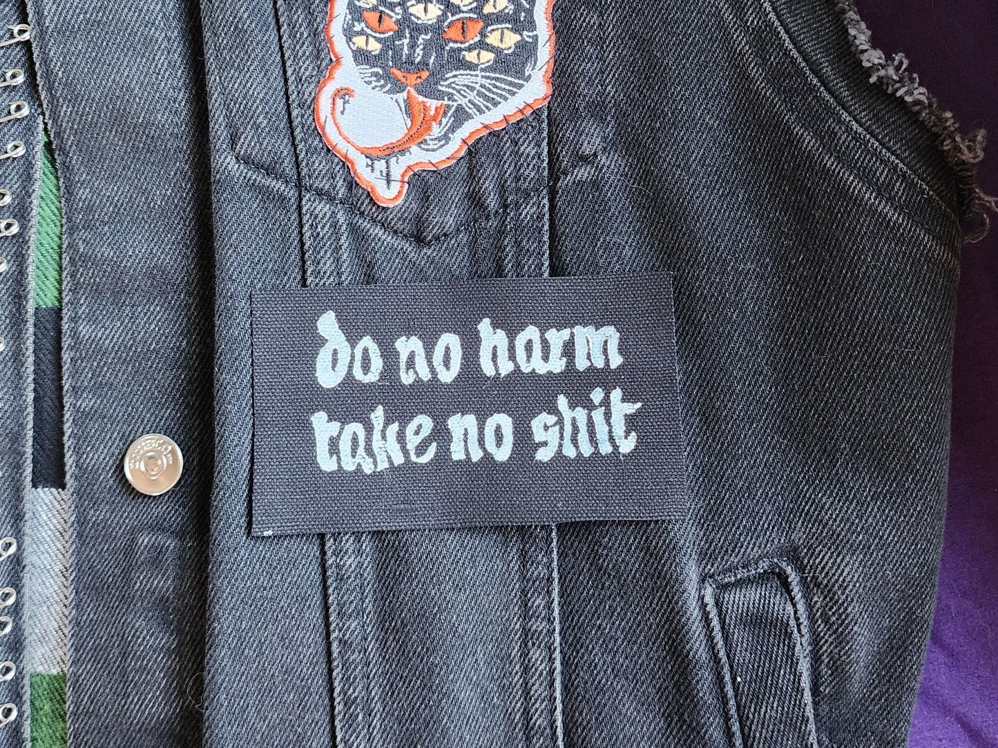 Black fabric patch with white Gothic-style text reading 'do no harm take no shit.' stacked on top of other similar patches. Displayed on a black denim background with another patch slightly in frame. Perfect for sewing onto jackets, bags, or other fabric surfaces
