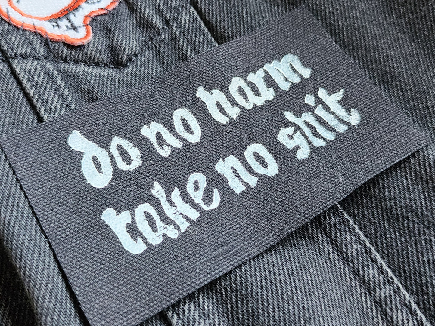 Black fabric patch with white Gothic-style text reading 'do no harm take no shit.' stacked on top of other similar patches. Displayed on a black denim background with another patch slightly in frame. Perfect for sewing onto jackets, bags, or other fabric surfaces