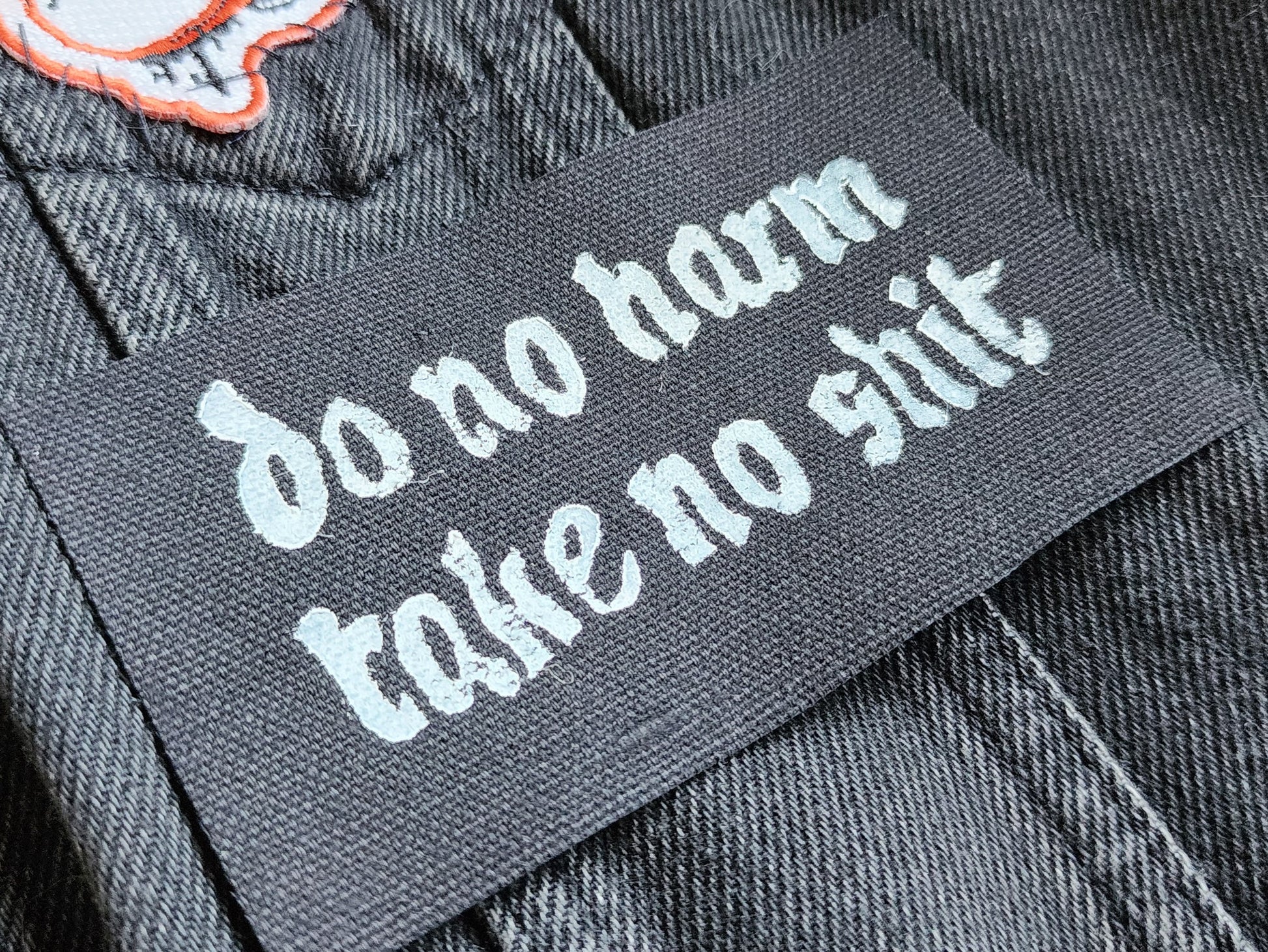 Black fabric patch with white Gothic-style text reading 'do no harm take no shit.' stacked on top of other similar patches. Displayed on a black denim background with another patch slightly in frame. Perfect for sewing onto jackets, bags, or other fabric surfaces