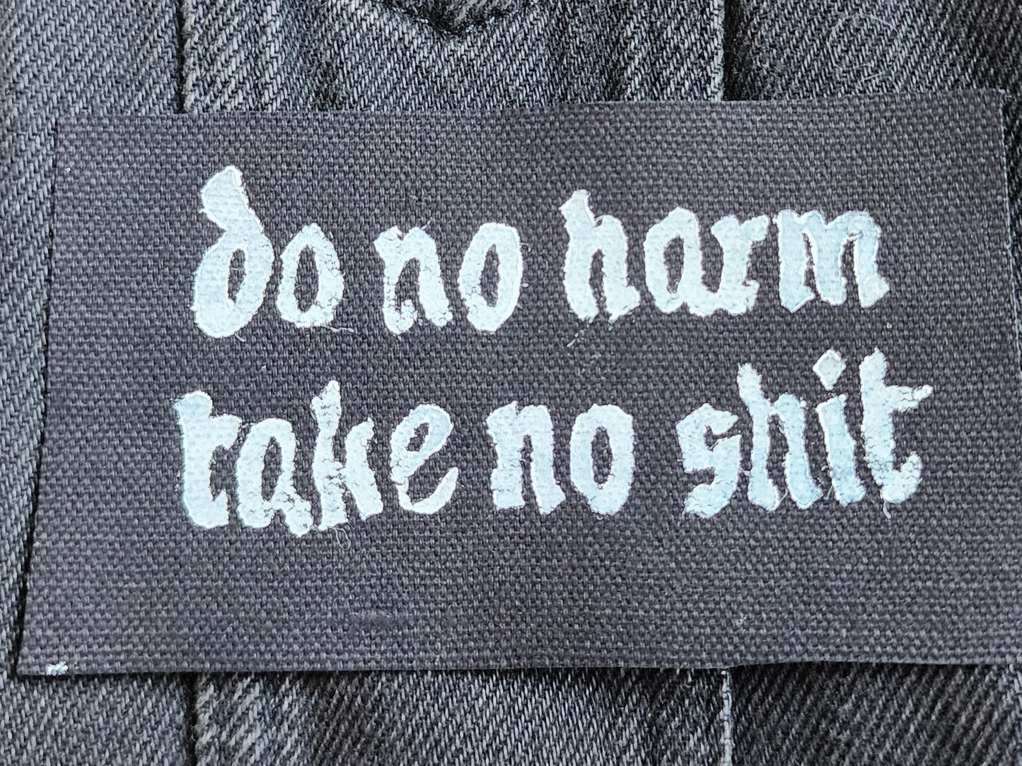 Black fabric patch with white Gothic-style text reading 'do no harm take no shit.' stacked on top of other similar patches. Displayed on a black denim background with another patch slightly in frame. Perfect for sewing onto jackets, bags, or other fabric surfaces