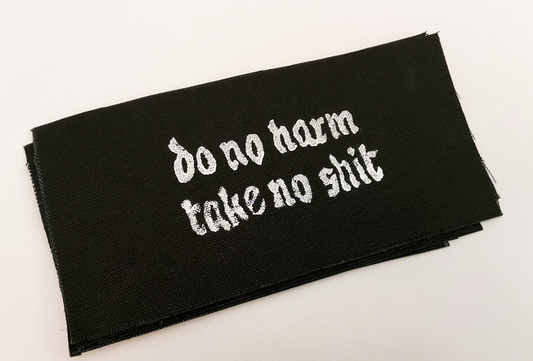 Black fabric patch with white Gothic-style text reading 'do no harm take no shit.' stacked on top of other similar patches. Displayed on a plain white background. Perfect for sewing onto jackets, bags, or other fabric surfaces