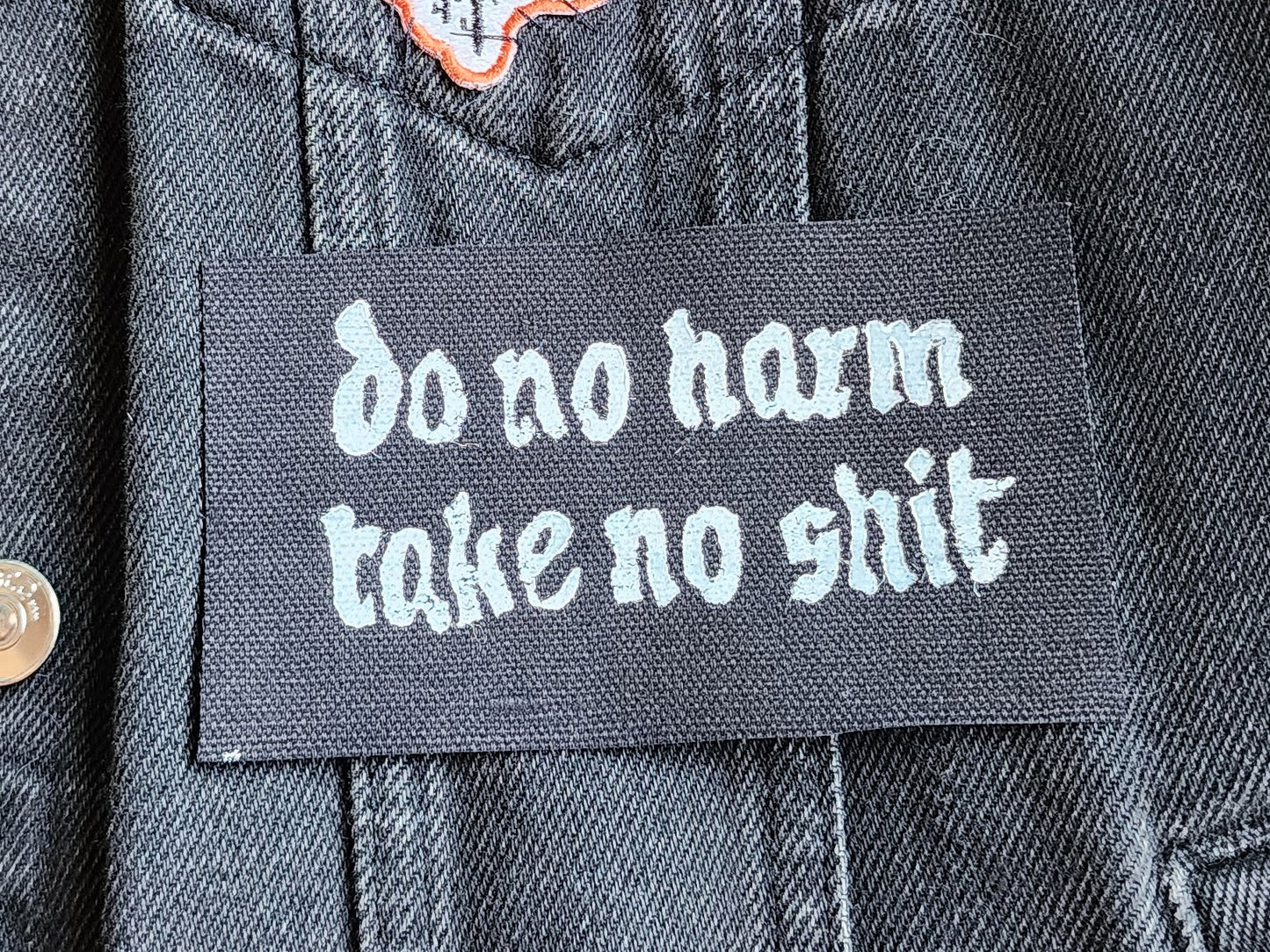 Black fabric patch with white Gothic-style text reading 'do no harm take no shit.' stacked on top of other similar patches. Displayed on a black denim background with another patch slightly in frame. Perfect for sewing onto jackets, bags, or other fabric surfaces