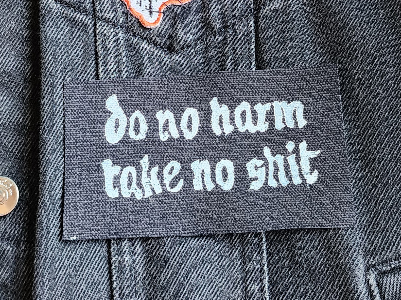Black fabric patch with white Gothic-style text reading 'do no harm take no shit.' stacked on top of other similar patches. Displayed on a black denim background with another patch slightly in frame. Perfect for sewing onto jackets, bags, or other fabric surfaces