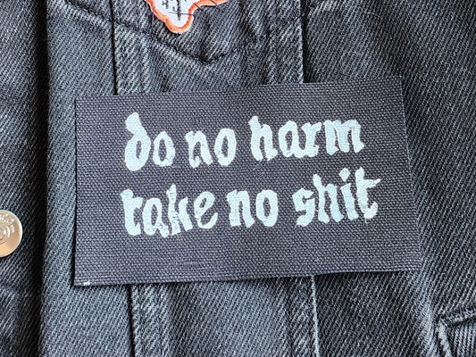Black fabric patch with white Gothic-style text reading 'do no harm take no shit.' stacked on top of other similar patches. Displayed on a black denim background with another patch slightly in frame. Perfect for sewing onto jackets, bags, or other fabric surfaces