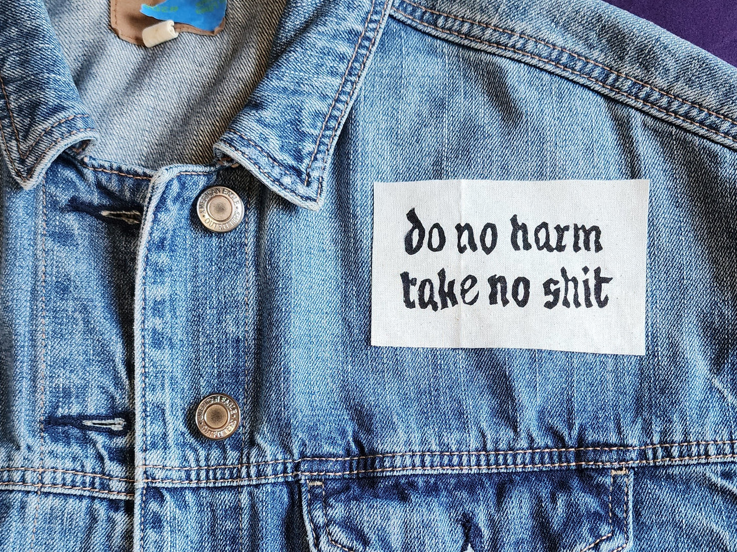 White fabric patch with white Gothic-style text reading 'do no harm take no shit.' laid on top of a blue denim jacket. Perfect for sewing onto jackets, bags, or other fabric surfaces