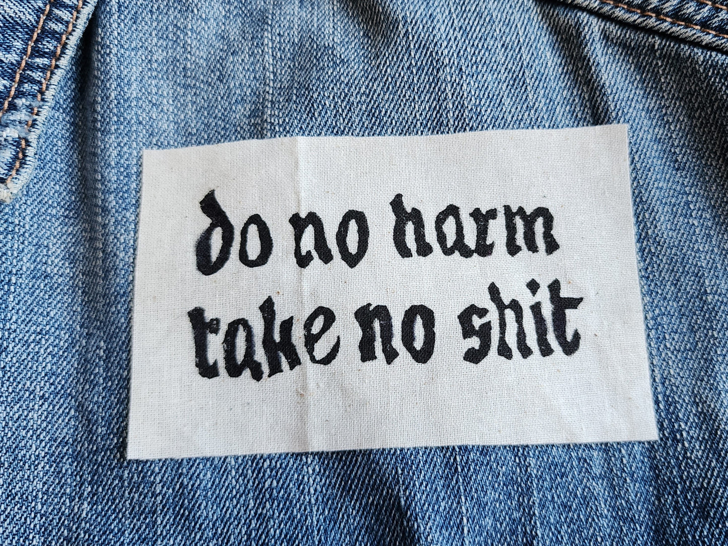 White fabric patch with white Gothic-style text reading 'do no harm take no shit.' laid on top of a blue denim jacket. Perfect for sewing onto jackets, bags, or other fabric surfaces