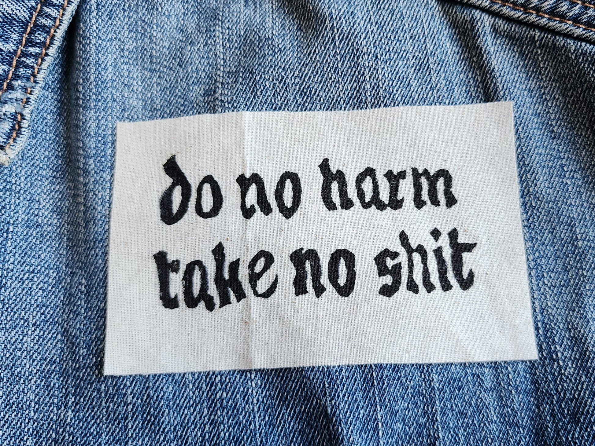 White fabric patch with white Gothic-style text reading 'do no harm take no shit.' laid on top of a blue denim jacket. Perfect for sewing onto jackets, bags, or other fabric surfaces