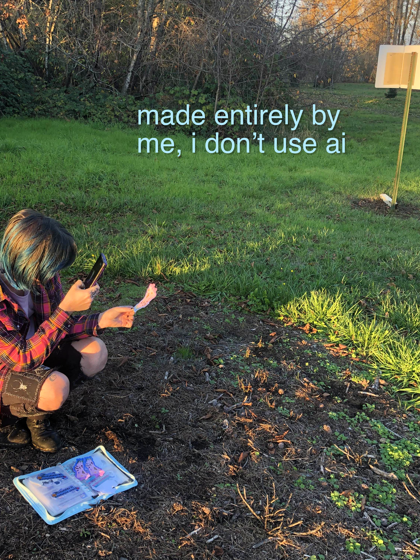a person with blue hair squats outdoors, taking a photo of a colorful sticker, with a sketchbook on the ground next o them. text reads in the top right reads "made entirely by me, i don't use ai" in light blue. the person has blue hair, visible tattoos, and patch pants. they are outdoors in a field during golden hour.