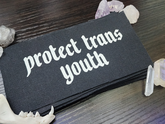 A black fabric patch with the phrase 'Protect Trans Youth' printed in bold, white Gothic-style lettering. The patch is stacked neatly with others on a dark wooden surface. Surrounding the patch are decorative elements, including clear and purple crystals, a small animal jawbone, and rough stones, adding a mystical and earthy aesthetic.