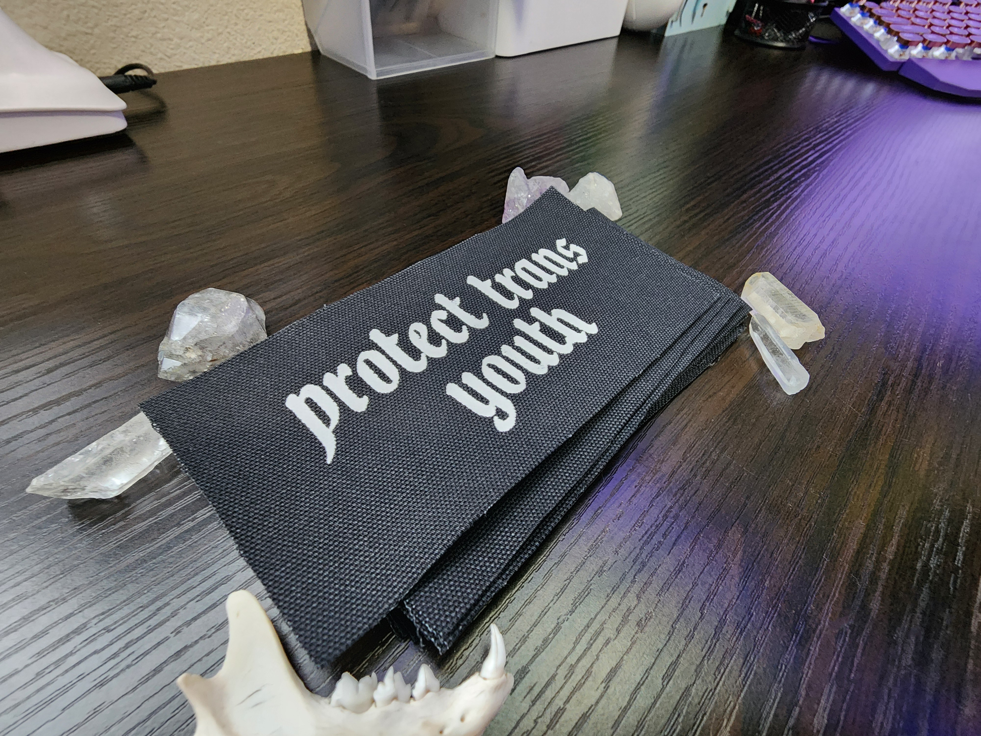 A black fabric patch with the phrase 'Protect Trans Youth' printed in bold, white Gothic-style lettering. The patch is stacked neatly with others on a dark wooden surface. Surrounding the patch are decorative elements, including clear and purple crystals, a small animal jawbone, and rough stones, adding a mystical and earthy aesthetic.