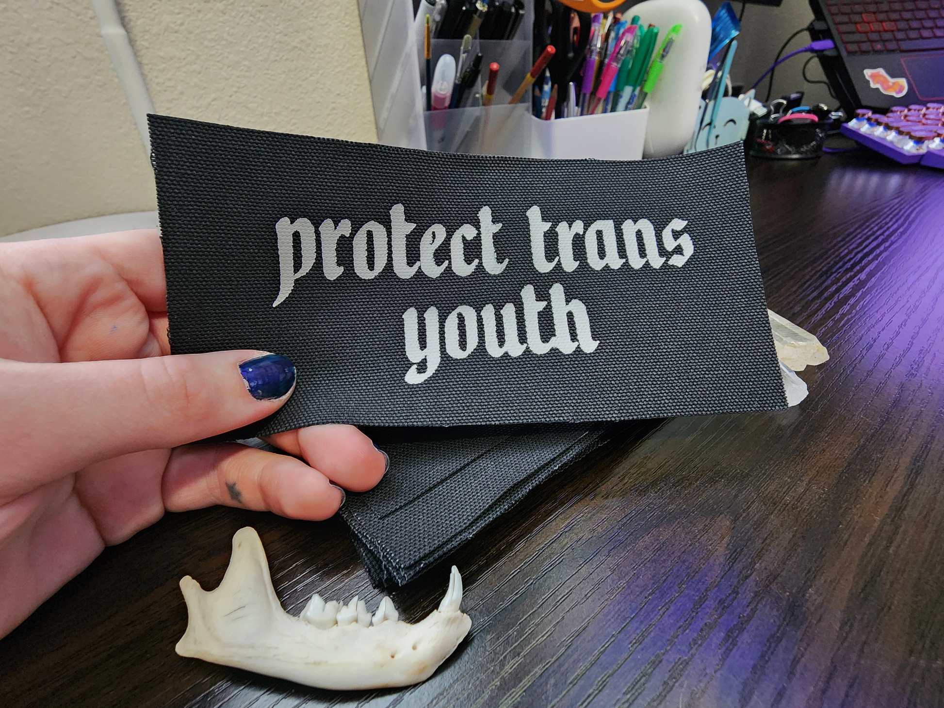 A black fabric patch with the phrase 'Protect Trans Youth' printed in bold, white Gothic-style lettering. The patch is stacked neatly with others on a dark wooden surface. Surrounding the patch are decorative elements, including clear and purple crystals, a small animal jawbone, and rough stones, adding a mystical and earthy aesthetic.