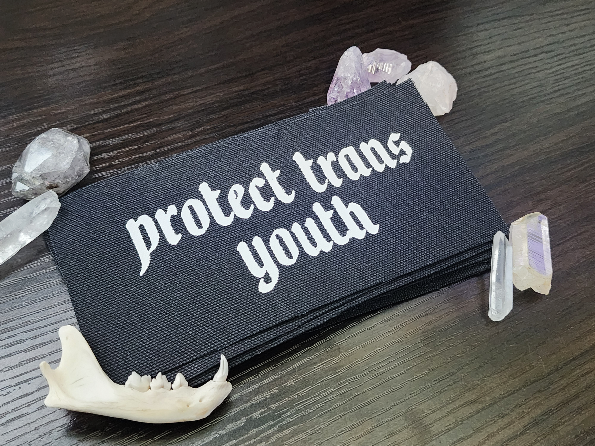 A black fabric patch with the phrase 'Protect Trans Youth' printed in bold, white Gothic-style lettering. The patch is stacked neatly with others on a dark wooden surface. Surrounding the patch are decorative elements, including clear and purple crystals, a small animal jawbone, and rough stones, adding a mystical and earthy aesthetic.